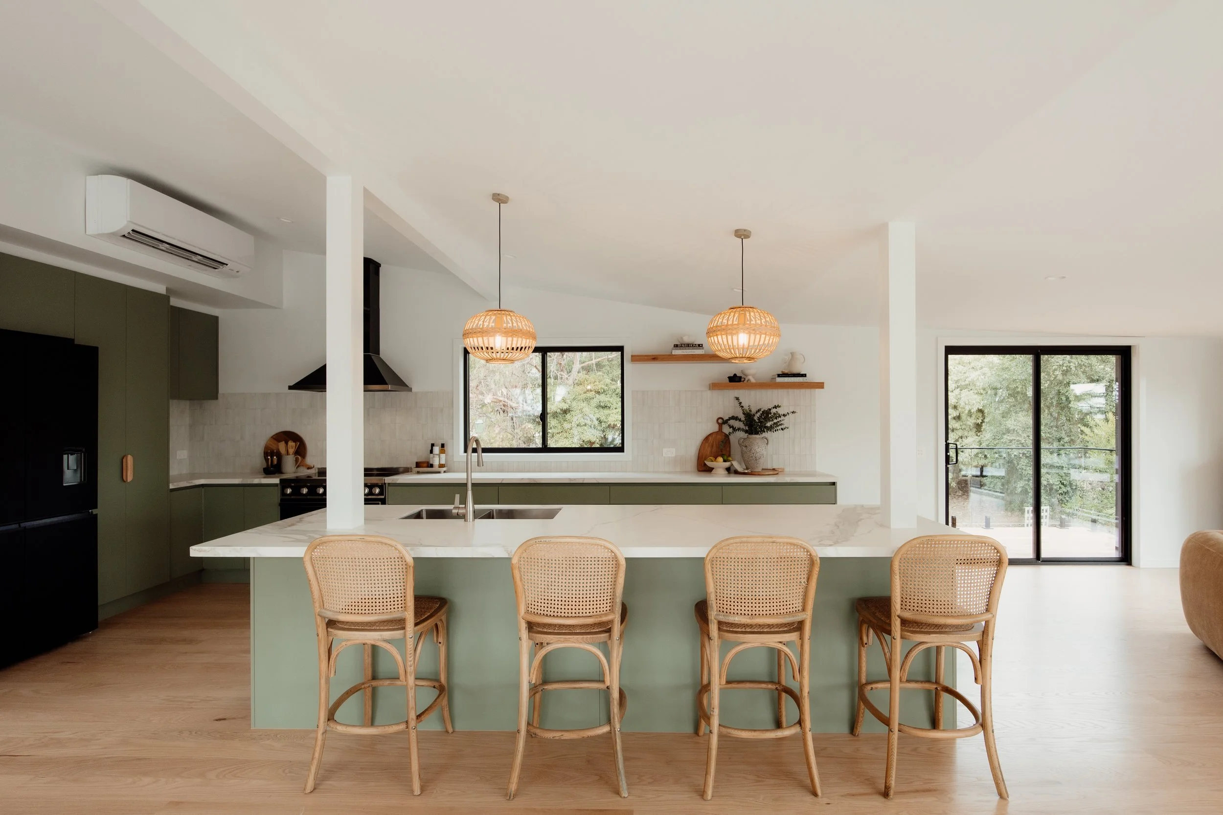 Modern open-concept kitchen with green cabinets, white countertops, wooden bar stools, pendant lights, large windows, and sliding glass doors overlooking a lush outdoor area.