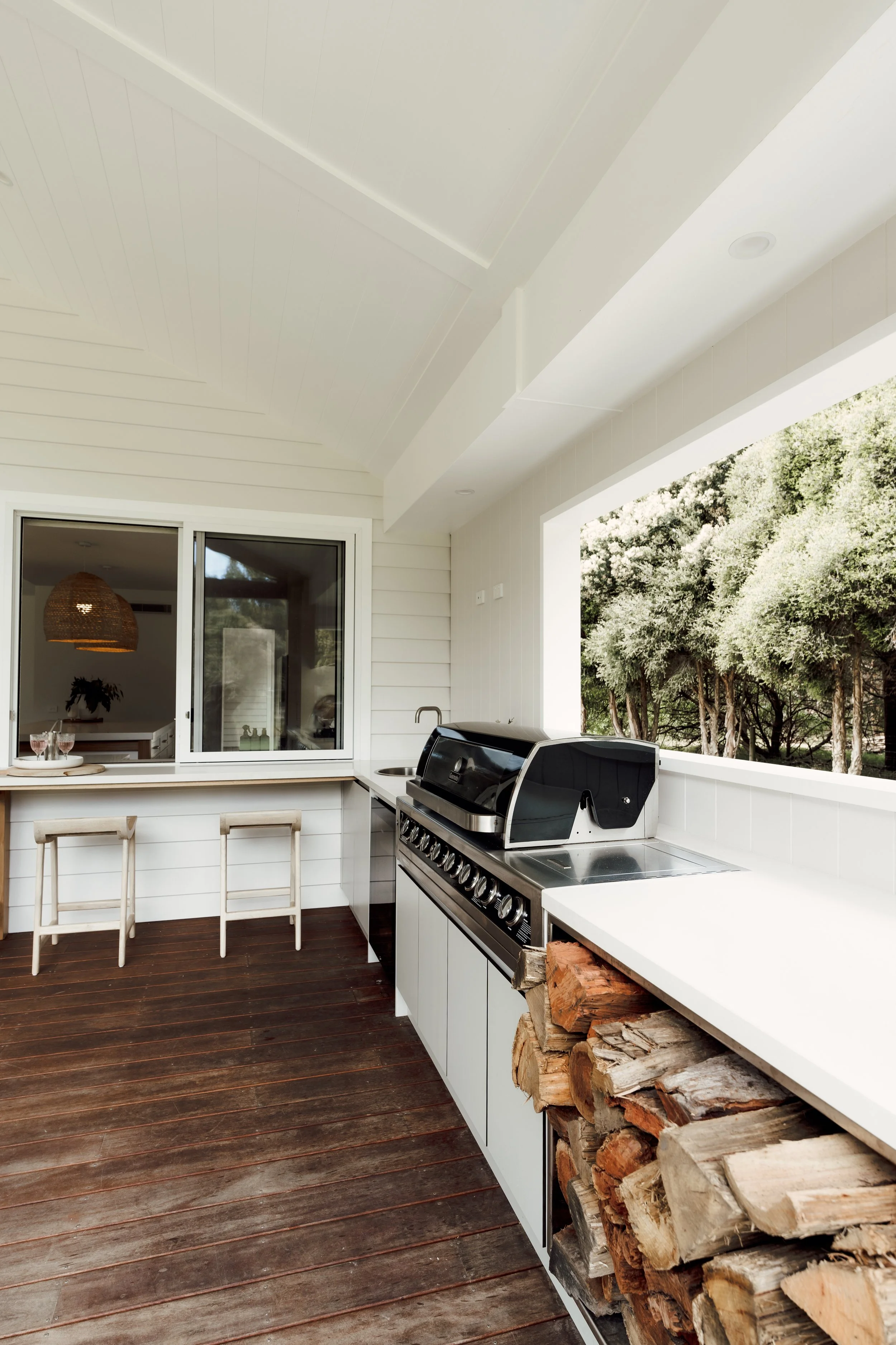 Outdoor kitchen with a stainless steel grill, white cabinetry, a wood pile beneath the counter, and a large window showing green trees outside.
