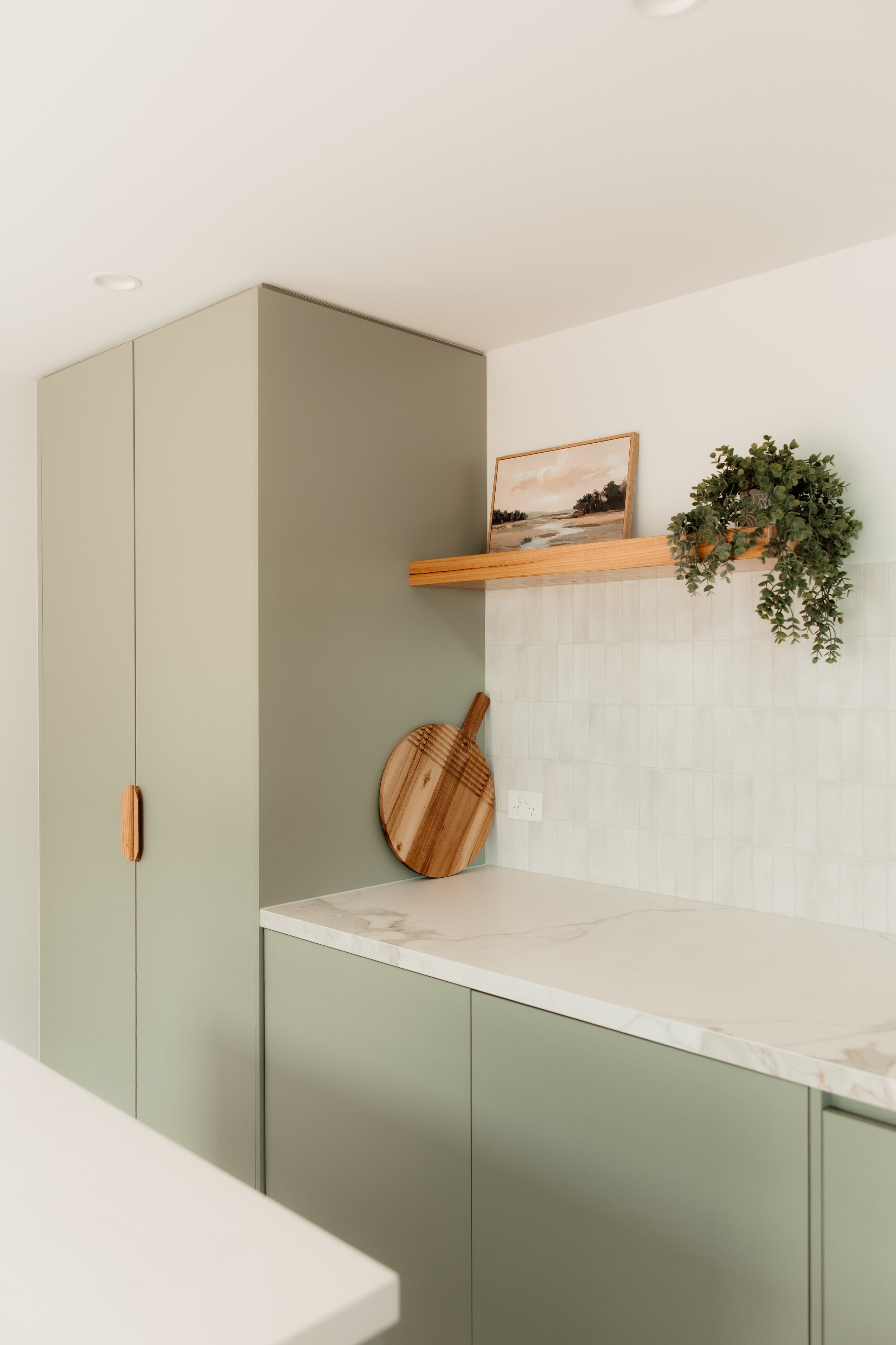 A minimal modern kitchen corner with green cabinets, white marble countertop, a floating wooden shelf with a framed landscape painting and a potted plant, and a round wooden cutting board hanging on the wall.