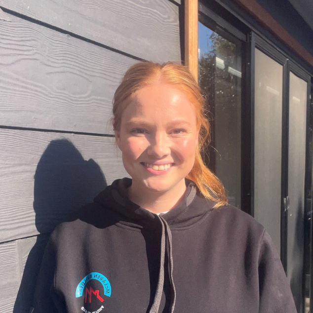 A young woman with red hair in a ponytail smiling outdoors in front of a gray wooden wall and a glass door, wearing a black hoodie with a logo on it.