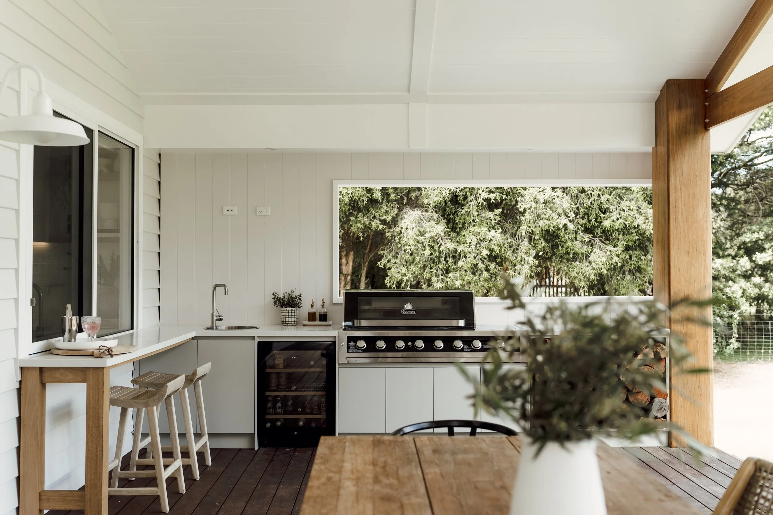 A modern outdoor kitchen area with white cabinetry, a black oven, and a large window with a view of green trees. There is a wooden dining table and chairs, and some potted plants on the counter.