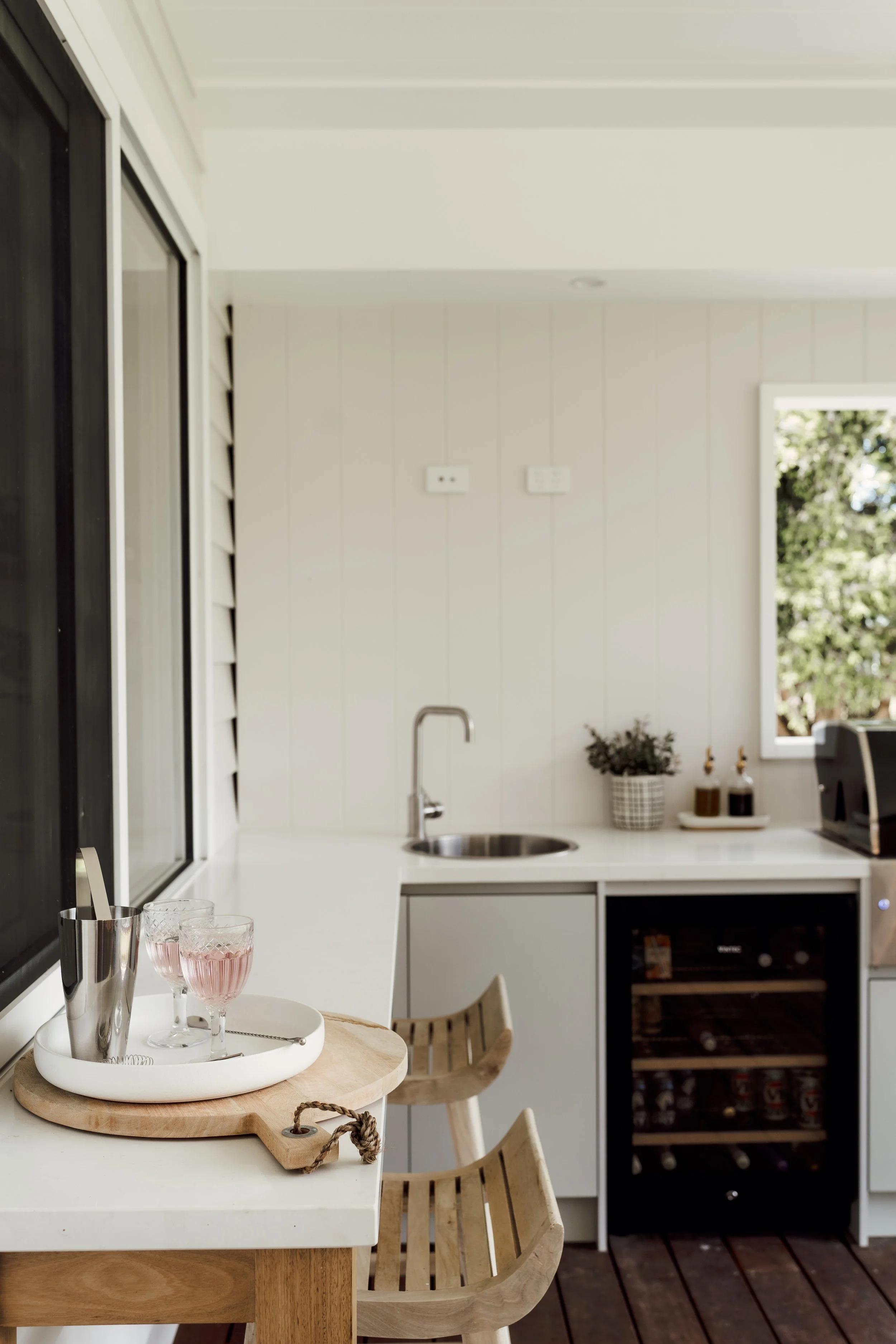 Modern kitchen with white cabinets, wood accents, and a small dining table with glasses and a tray. There is a window with trees outside and a door with black trim.