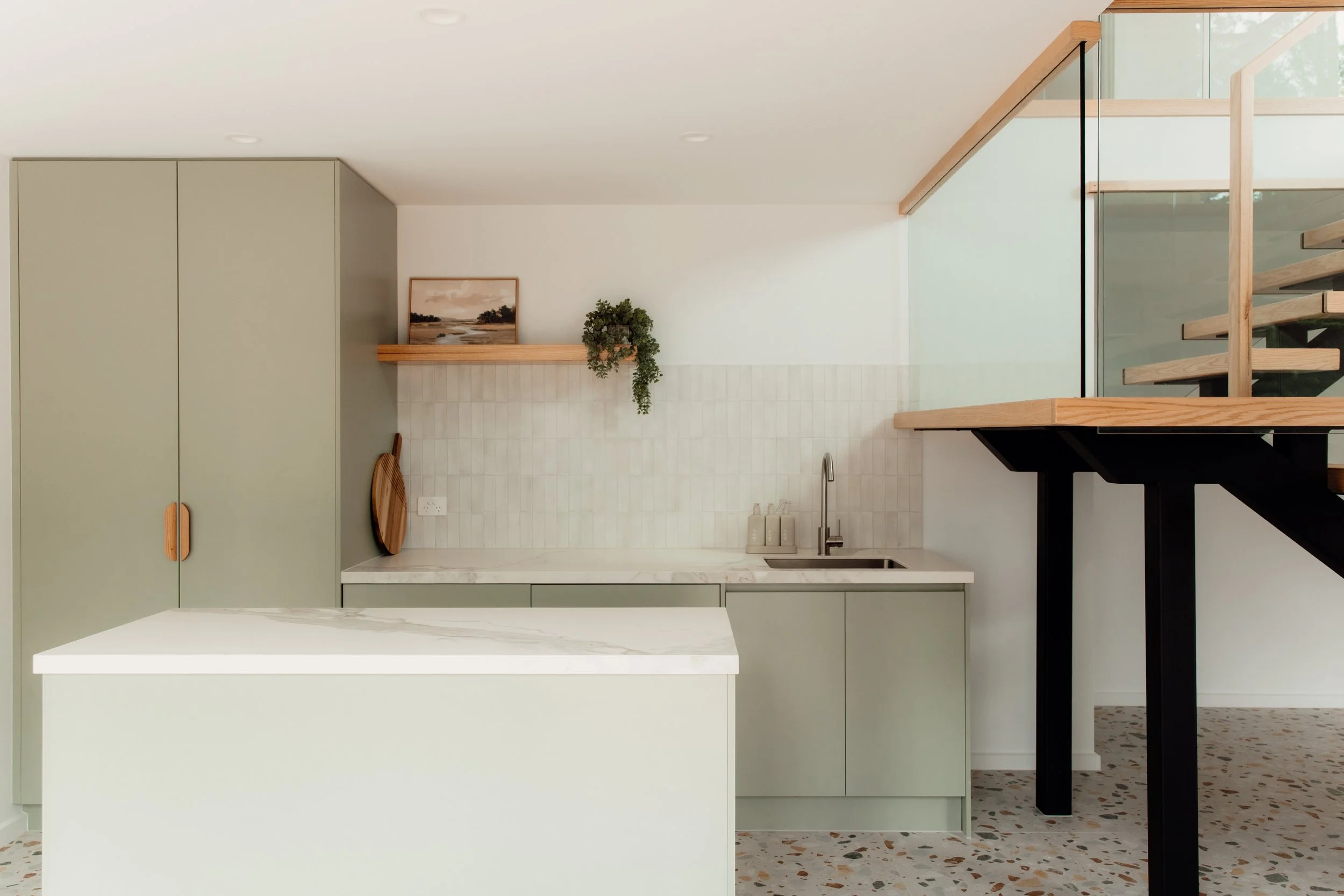 Modern kitchen with muted green cabinetry, white marble countertops, a backsplash with white tiles, and a small wooden shelf with a framed landscape painting and a hanging green plant. Part of a staircase with wooden steps and a black support is visi