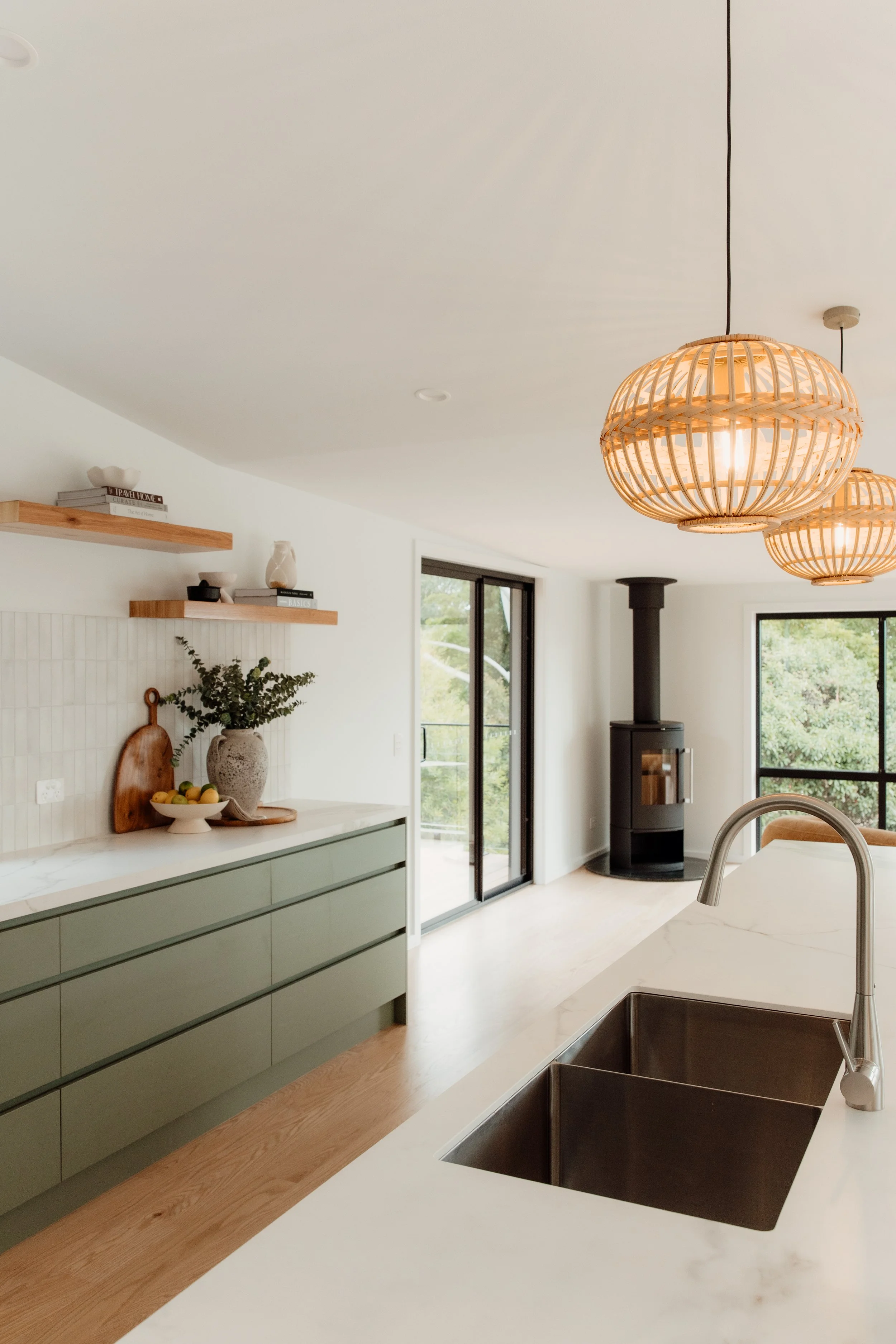 Modern kitchen with white countertops, green cabinets, wooden shelves, hanging wicker light fixtures, a black wood stove, and large windows with a view of greenery.