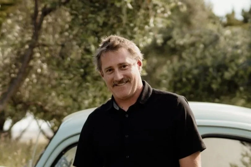 A man with a mustache smiling outdoors, standing next to a vintage white car with green trees in the background.