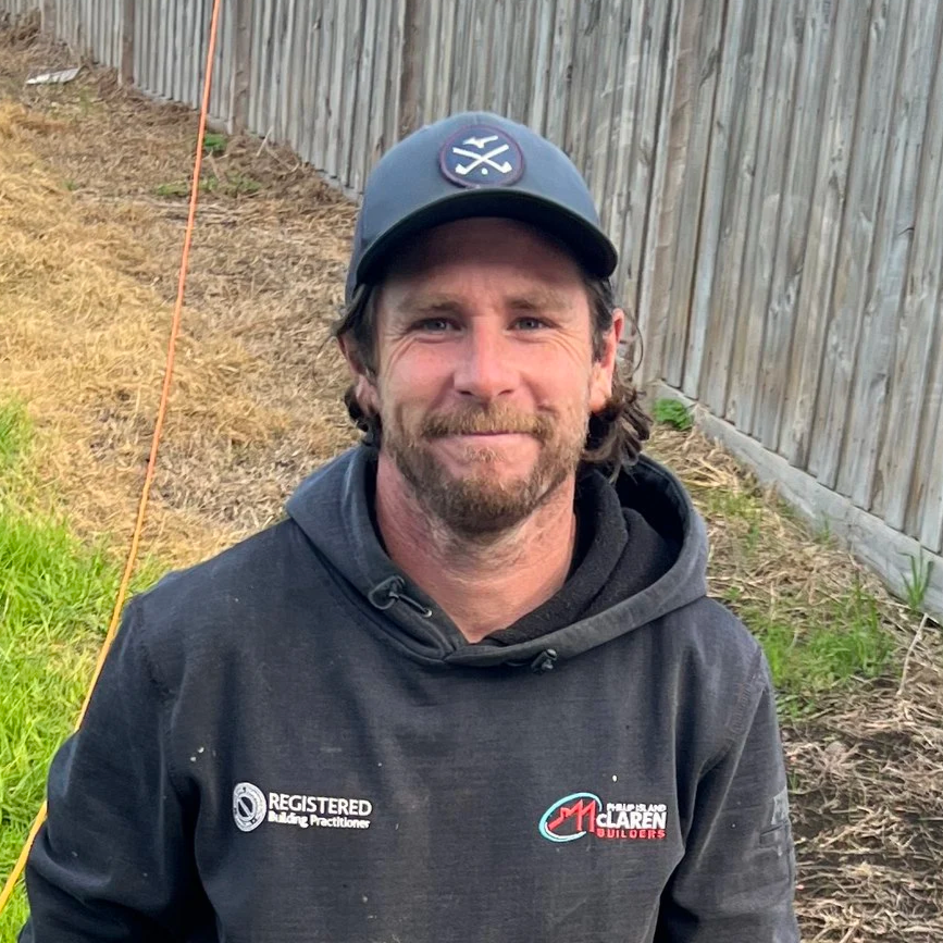 A smiling man wearing a black hoodie with logos, a black baseball cap, and standing outdoors beside a wooden fence with a patch of green grass and bare dirt.