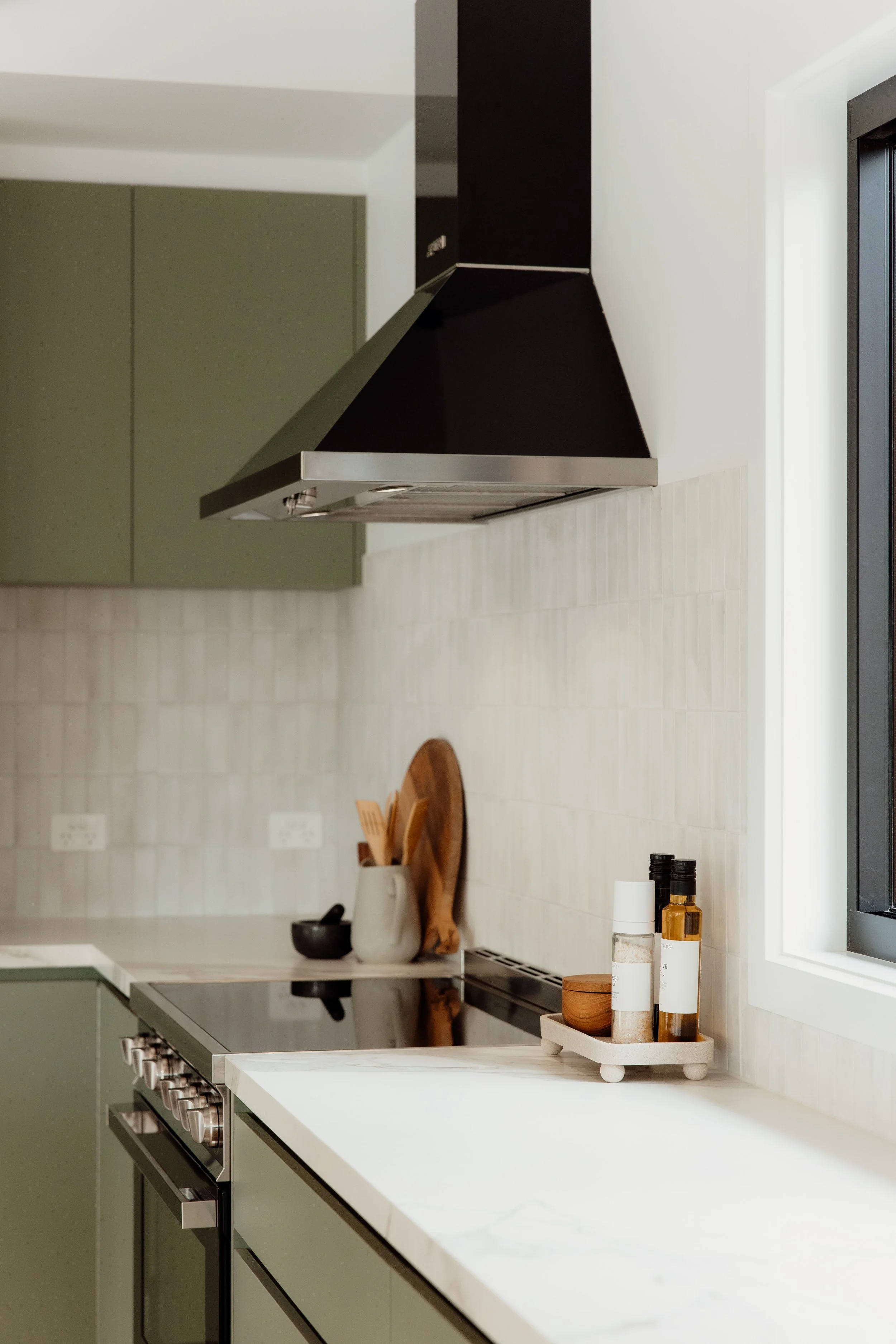 Modern kitchen with green cabinets, stainless steel stove and hood, white countertop, and small decorative items including jars and a wooden cutting board.