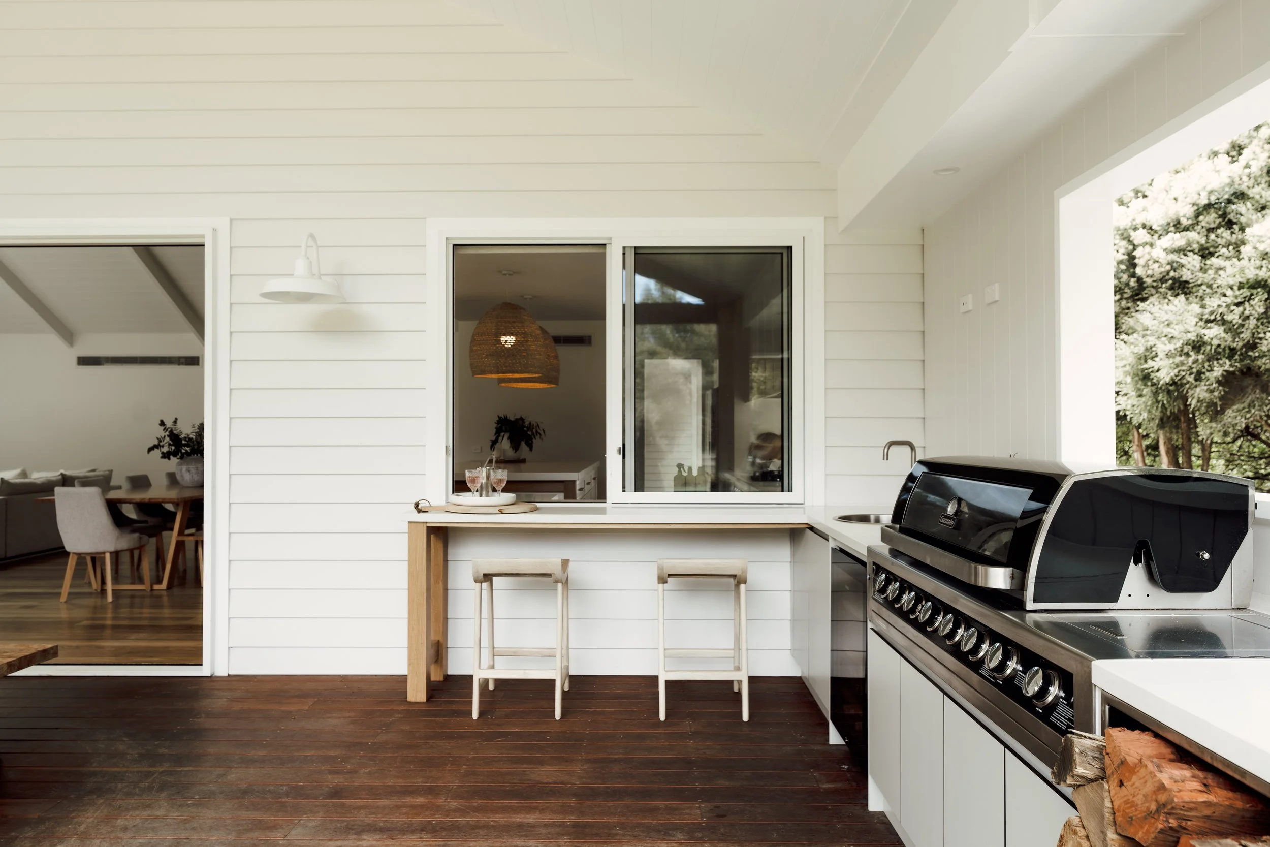 Outdoor kitchen with a gas grill, white cabinets, wooden countertops, and two white bar stools, overlooking trees to the right.