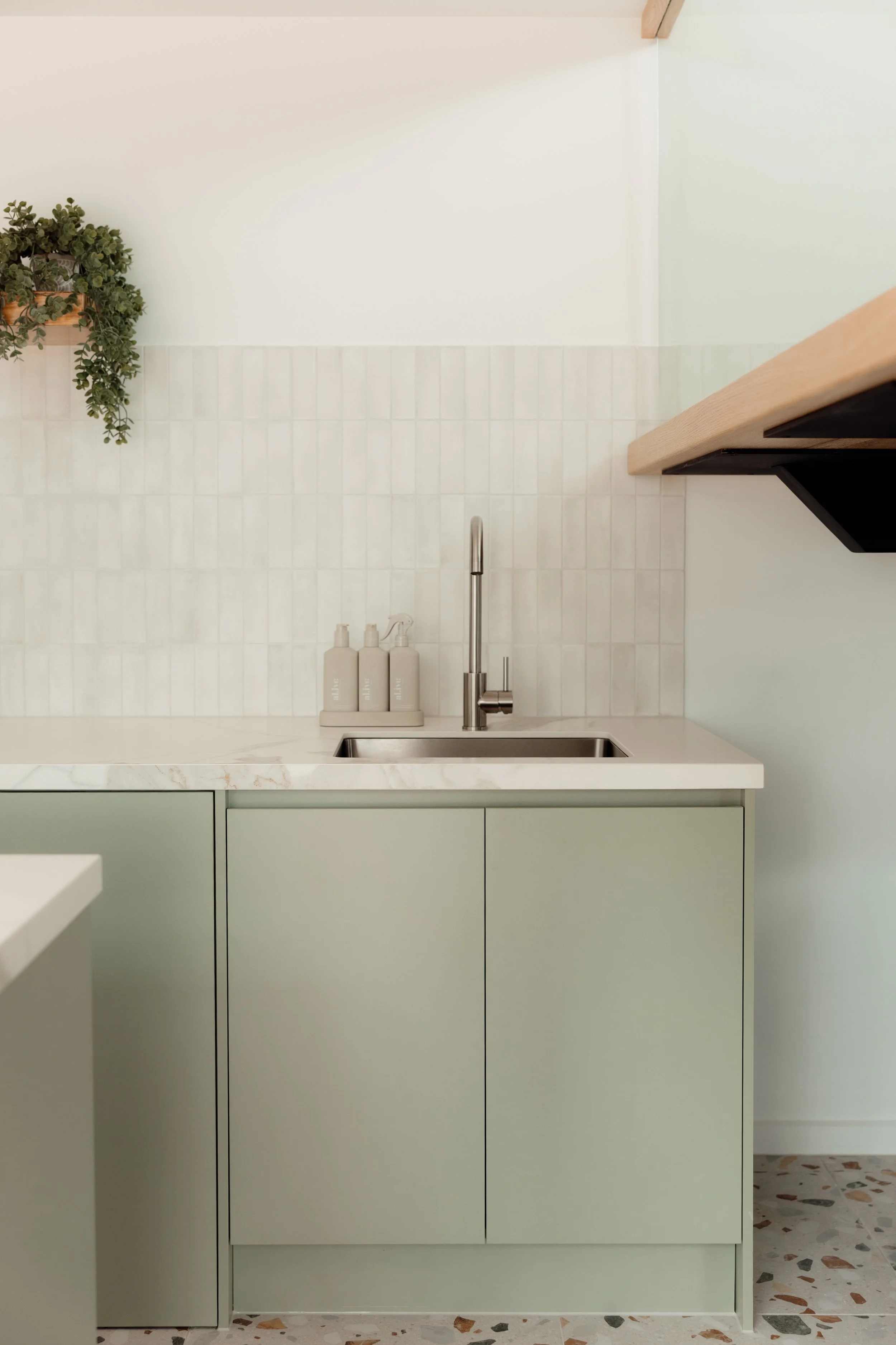 A minimalist kitchen with light green cabinets and a white marble countertop. There is a stainless steel sink with a modern faucet, three white bottles on the countertop, and a small wooden shelf with a green leafy plant hanging from it. The wall beh