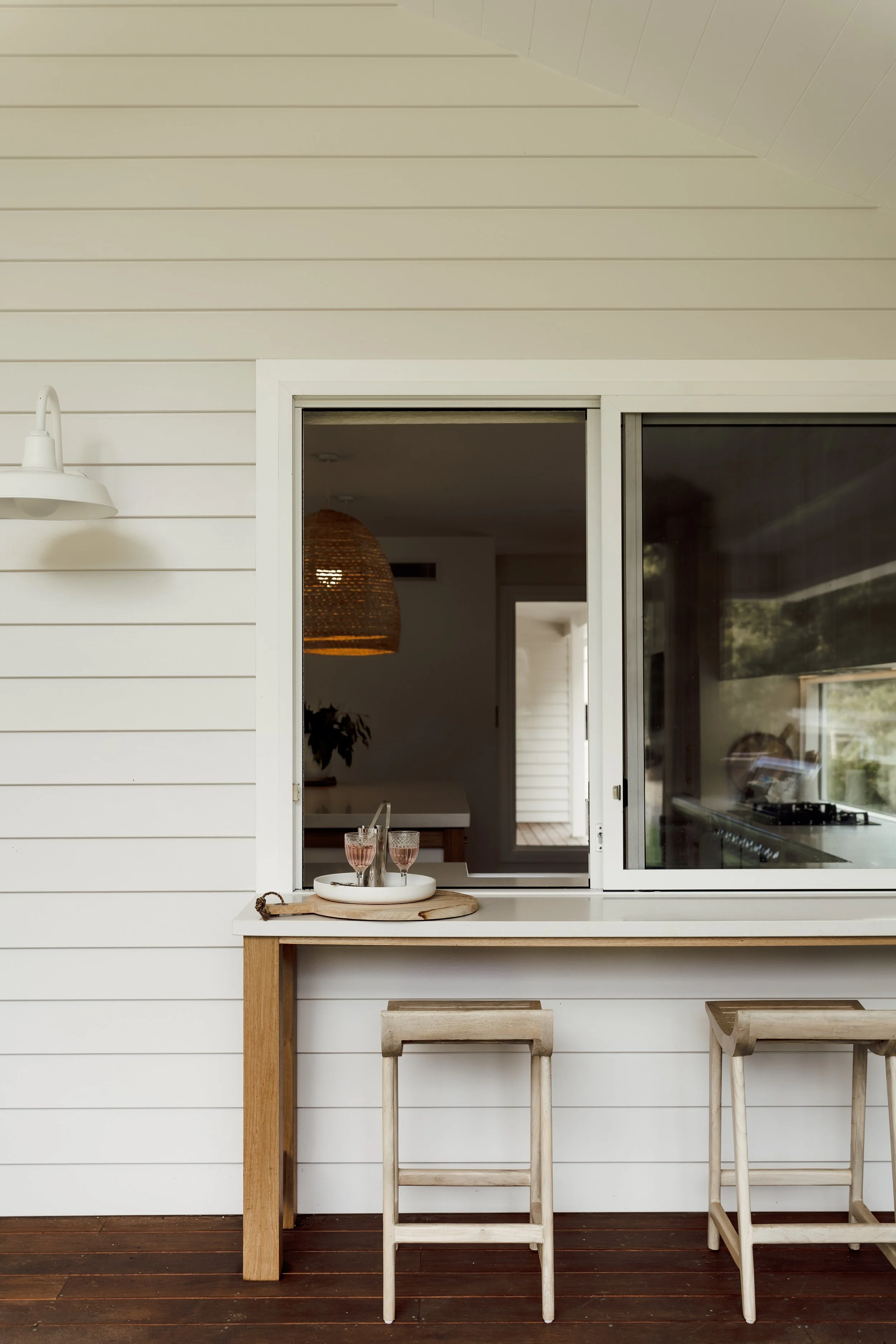 A kitchen window with a white s treatment over a countertop with two wooden barstools underneath. On the countertop, there is a small round tray with four wine glasses, two filled with pink drink, placed on a wooden slab. The background shows a hangi