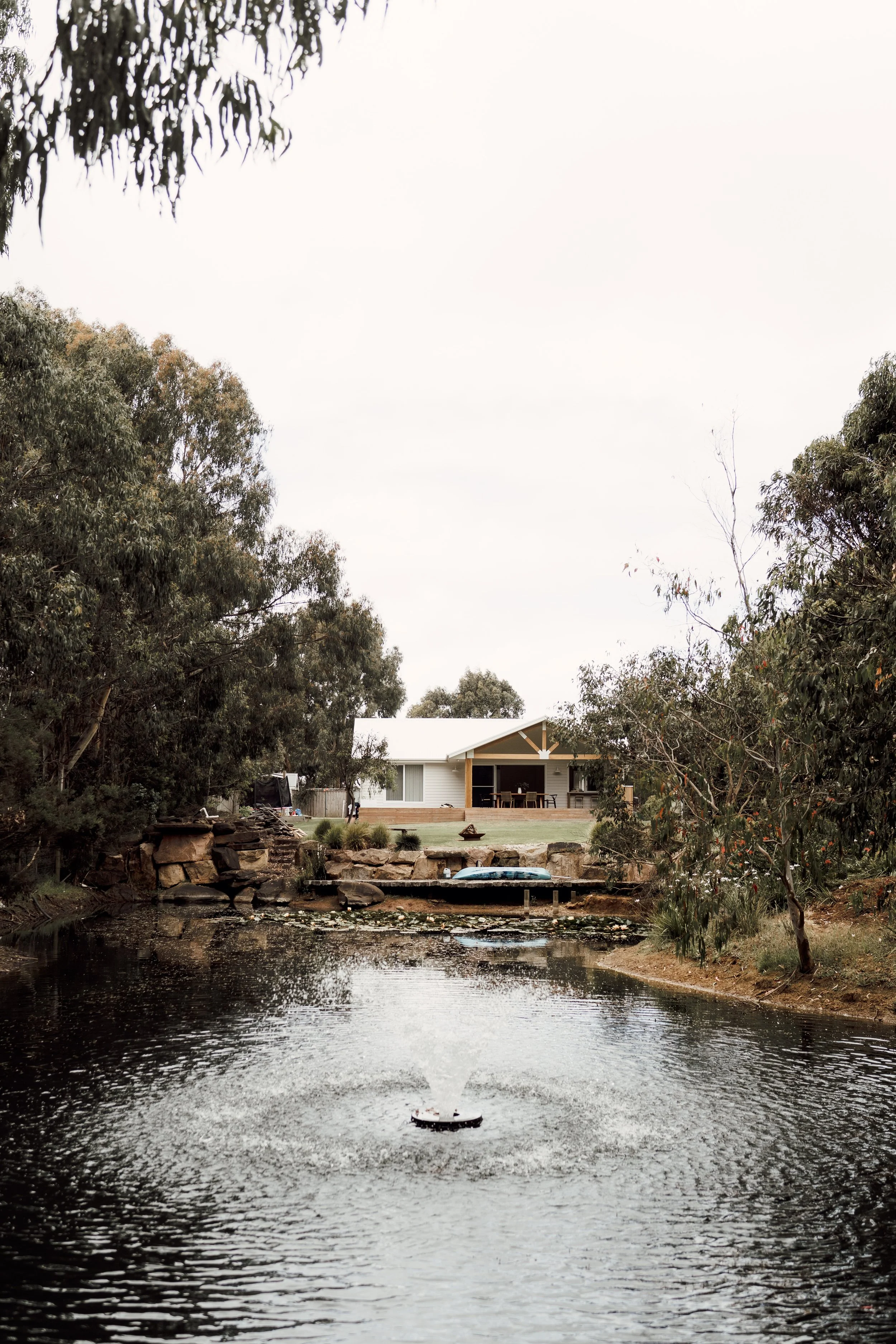 A house with a front porch, surrounded by trees, is situated along a pond with rocks and a dock. Overcast sky.