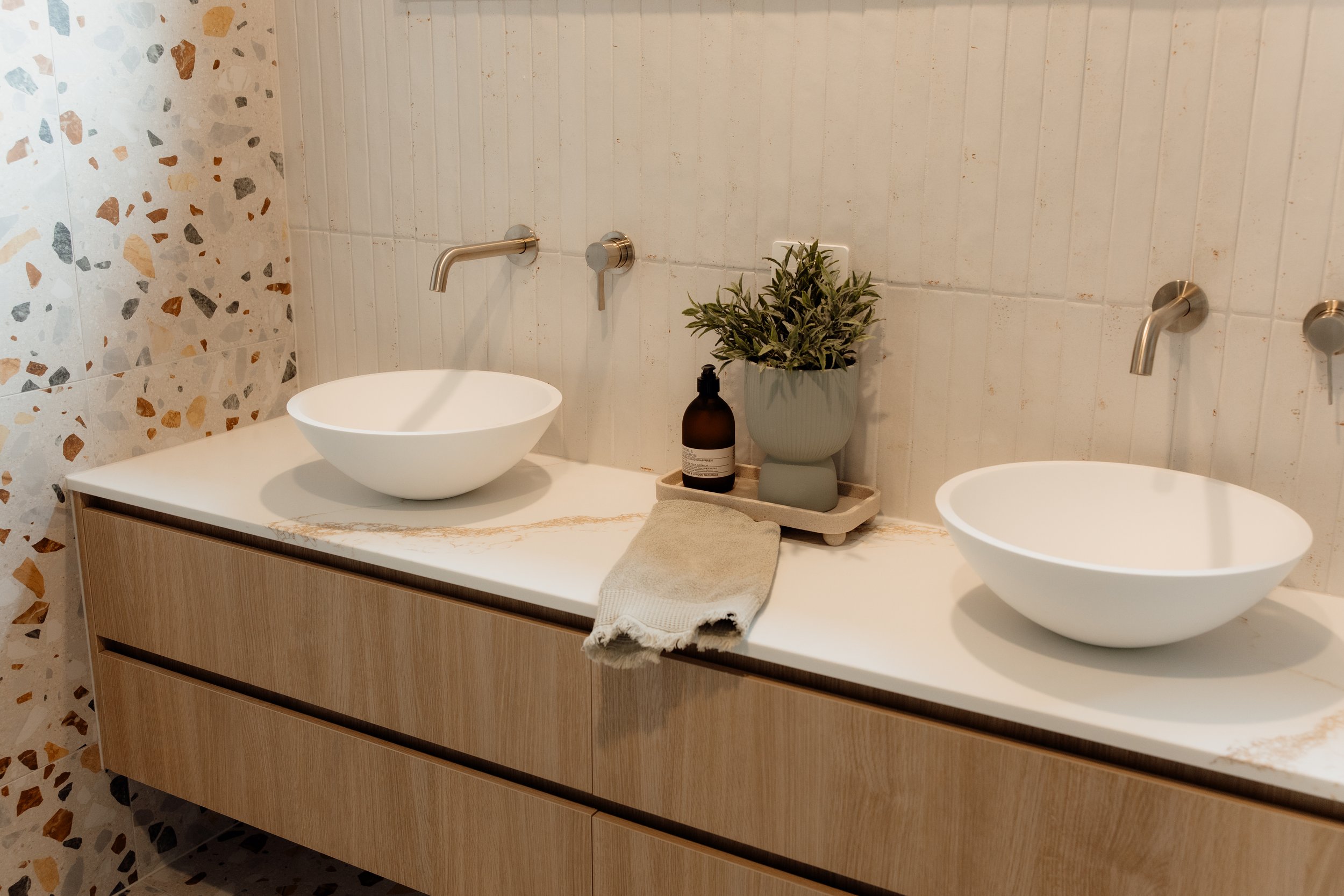 Bathroom with terrazzo wall tiles, a double vanity with white vessel sinks, a potted plant, soap dispenser, and a beige towel on the countertop.