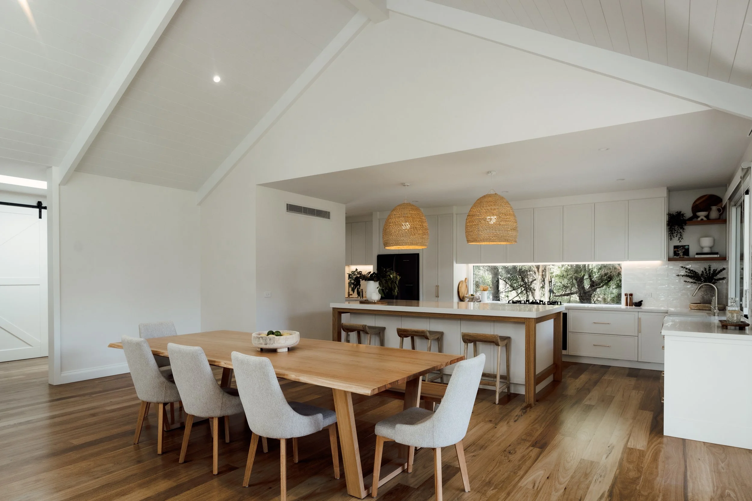 A bright, modern kitchen with a high, angled white ceiling with wood paneling. It features white cabinets, a large window with trees outside, and two woven pendant lights hanging over the kitchen island. There is a dining table with light gray chairs