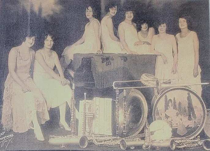 Group of women in white dresses sitting and standing around a piano, with musical instruments and a painting or photograph on a large drum in front.