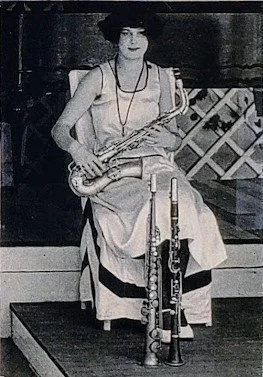 Woman sitting on a wooden platform holding a saxophone, wearing a long dress and a necklace, with clarinets at her feet, in vintage black-and-white style.