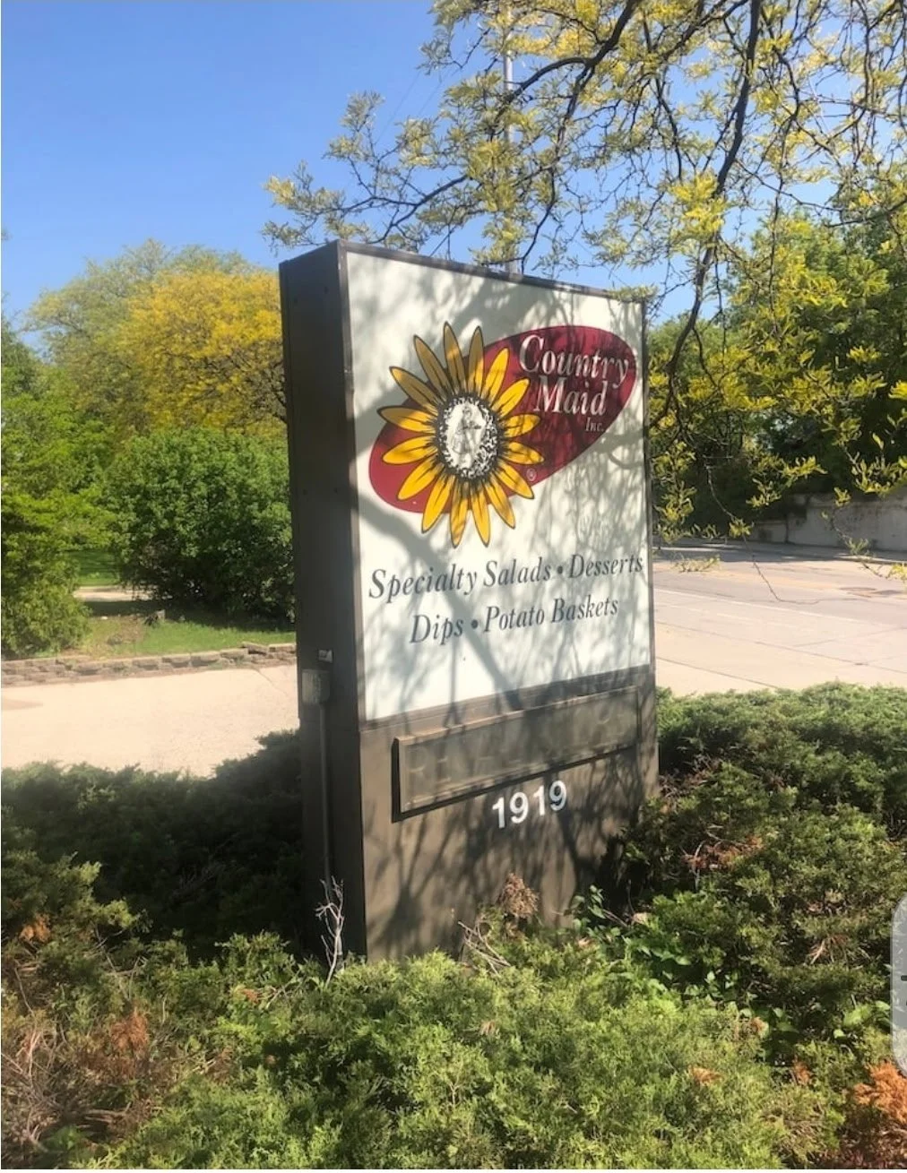 Sign for Country Maid Inc. advertising specialty salads, desserts, dips, and potato baskets, with the year 1919 at the bottom, surrounded by green shrubbery and trees in the background.