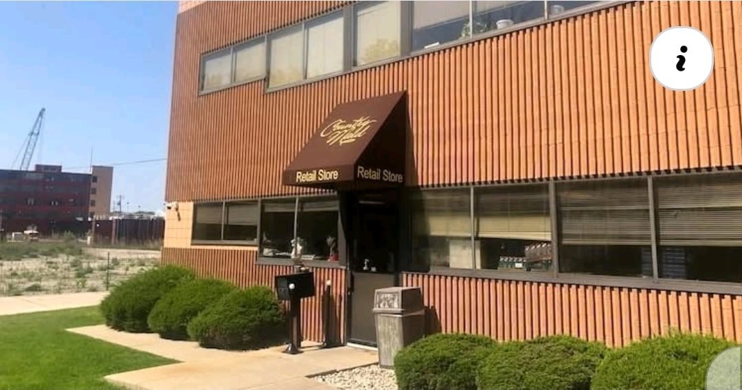 Exterior of a retail store with a brown wooden facade, black awning with gold lettering, and a pair of black and white signs labeled "Retail Store." There are bushes and a trash can near the entrance, with a sidewalk leading up to the door.