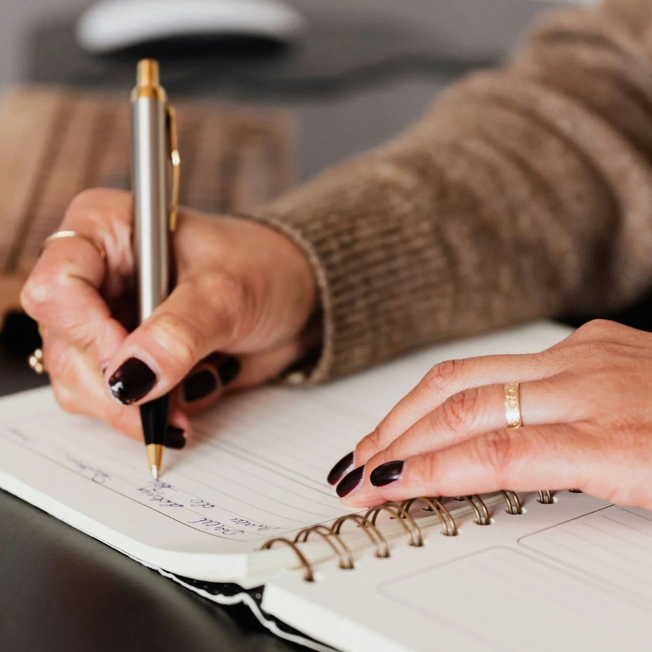 A person writing in a spiral notebook with a black and gold pen. The person has dark nail polish, is wearing rings, and a brown, striped sweater.