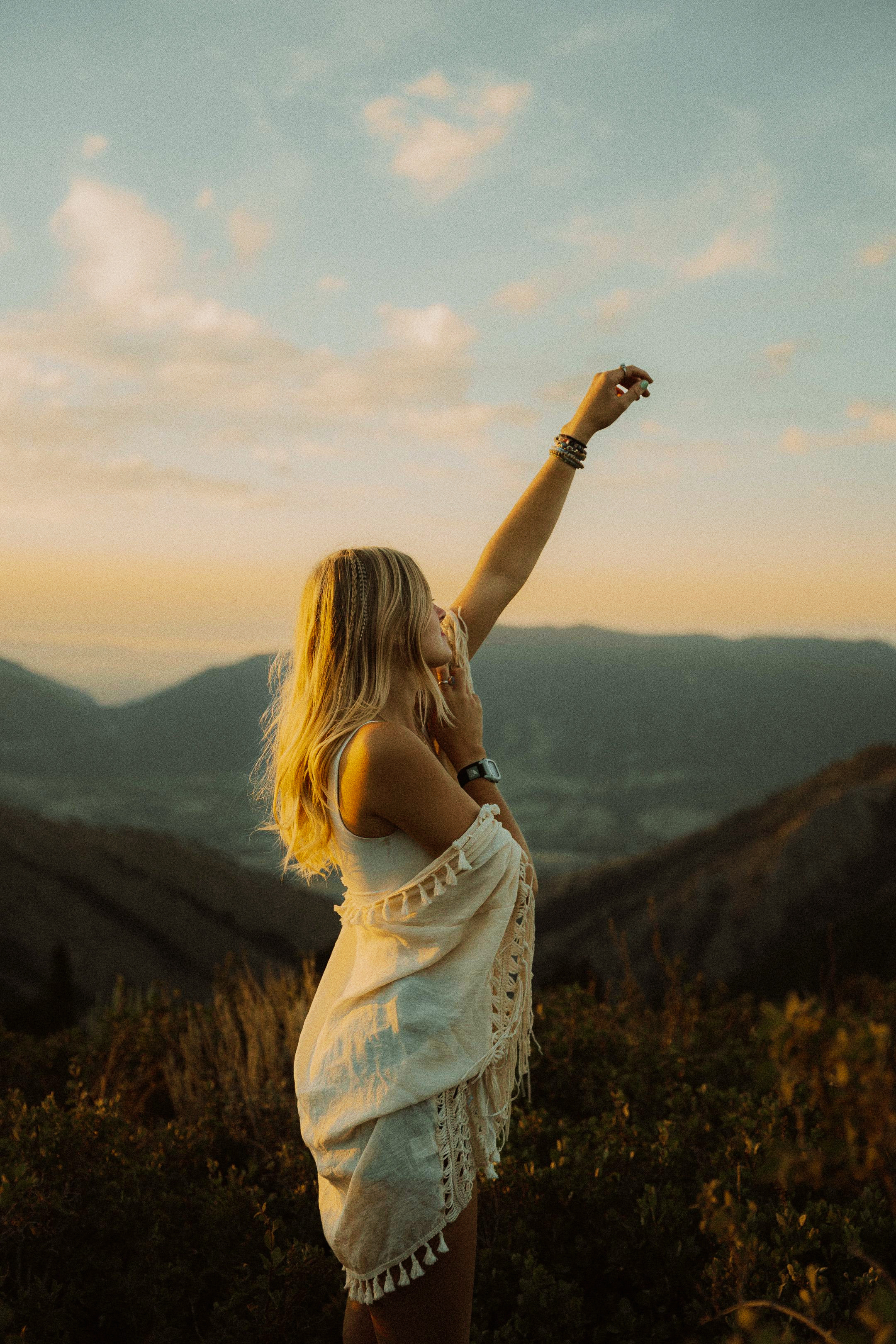 A woman with long blond hair standing outdoors on a hill at sunset, raising her right arm, with mountains in the background and a partly cloudy sky.