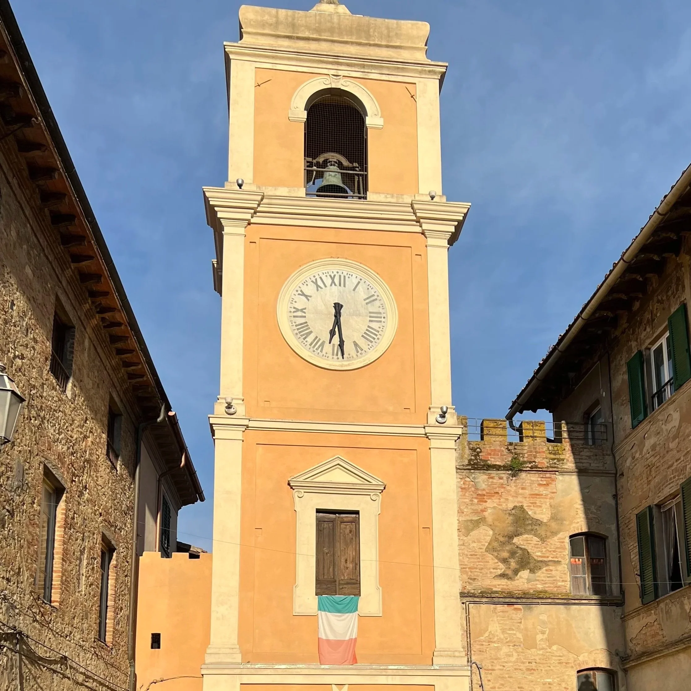 A tall clock tower with a bell at the top, displaying the time as 12:26, in a European city with narrow streets and old stone buildings. A flag is hanging from the building below the clock.