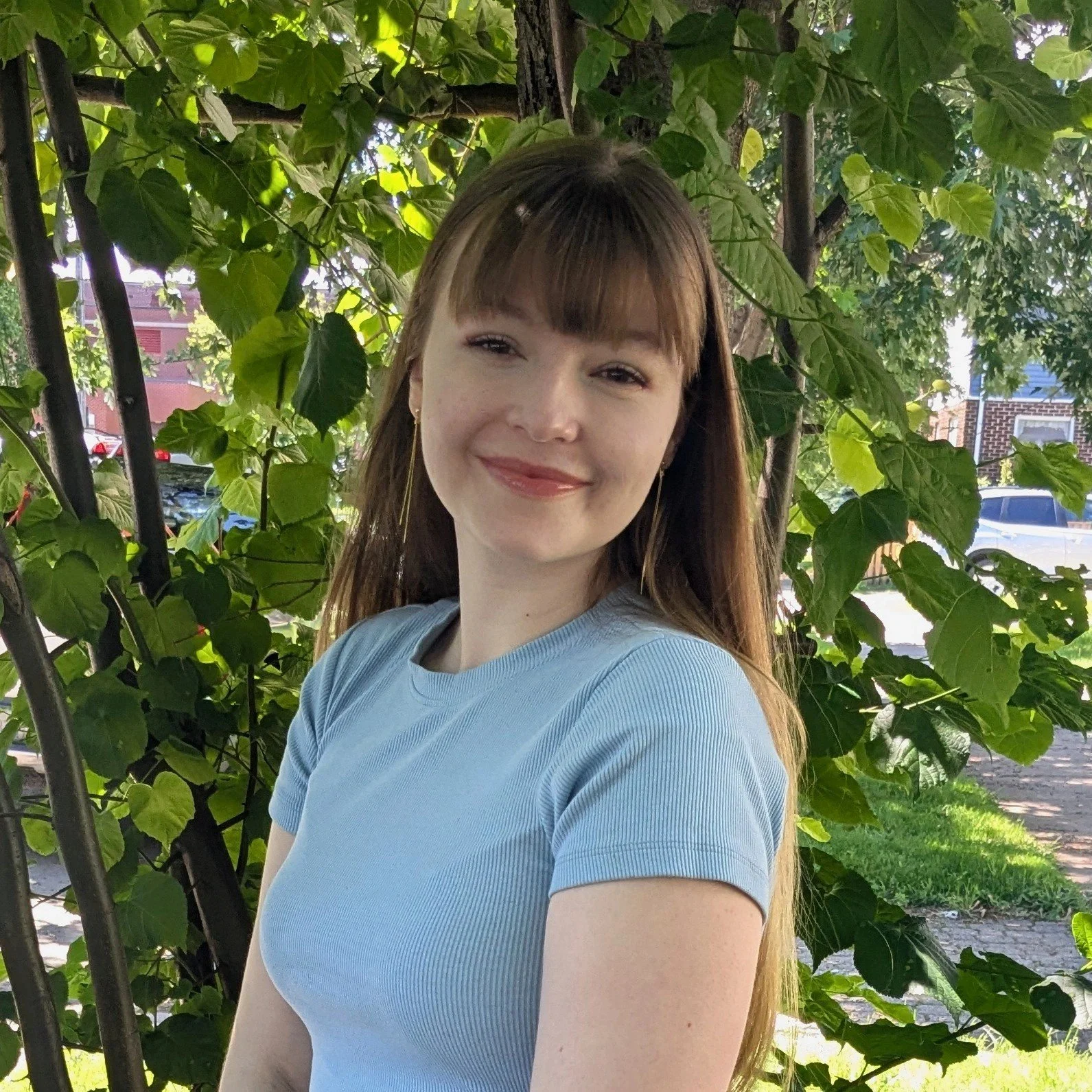 A young woman with long, light brown hair and bangs, wearing a light blue shirt, stands outdoors in front of green leafy plants and trees with sunlight filtering through.