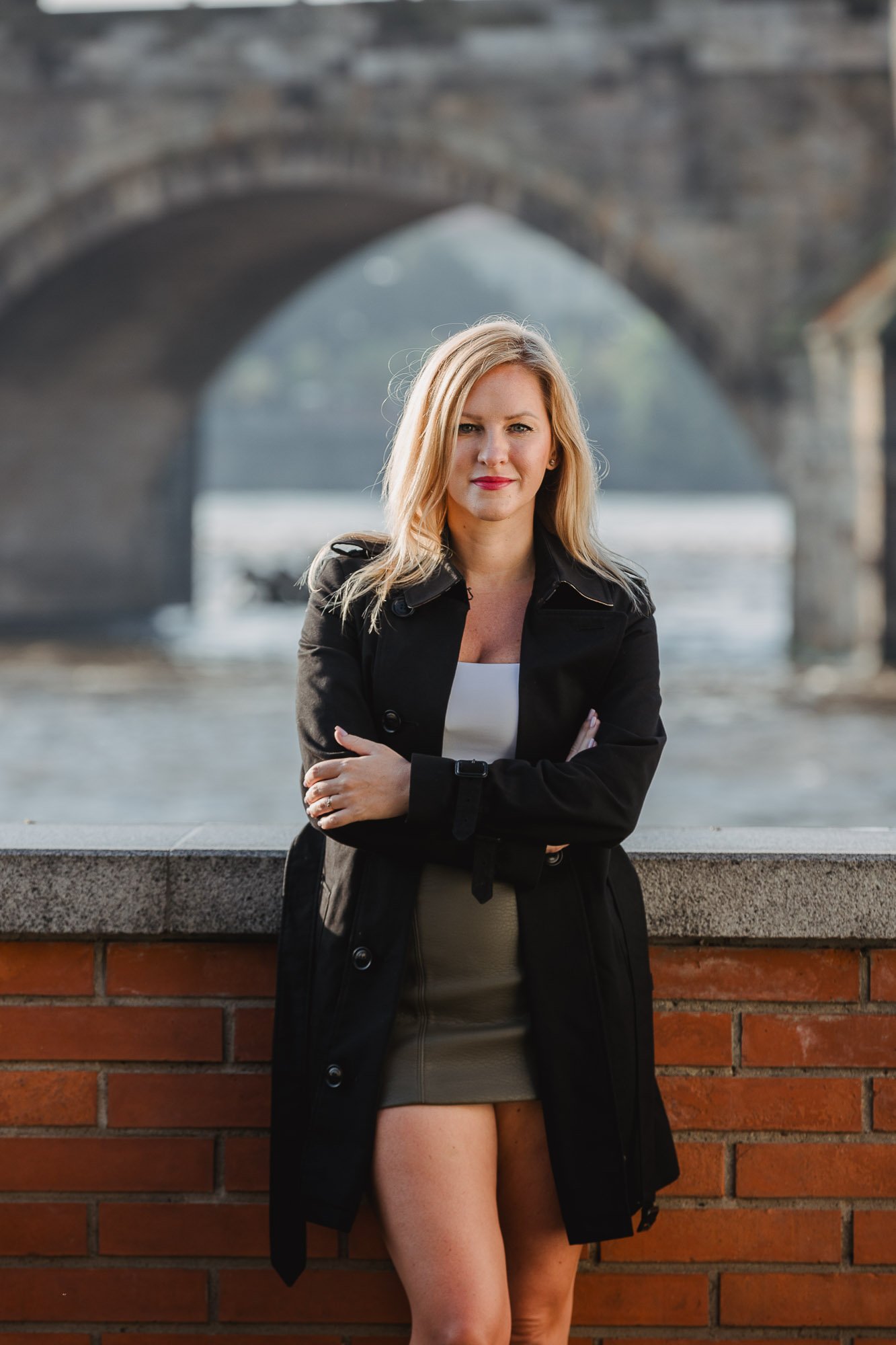 Woman standing outdoors in front of a brick wall and river, wearing a black coat, with arms crossed, under an arched stone bridge.