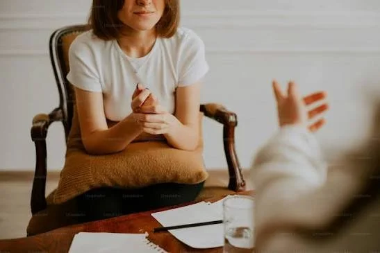 A woman sits at a table in an office or consultation setting, listening to someone who is speaking, with papers, a pen, and a glass of water on the table.