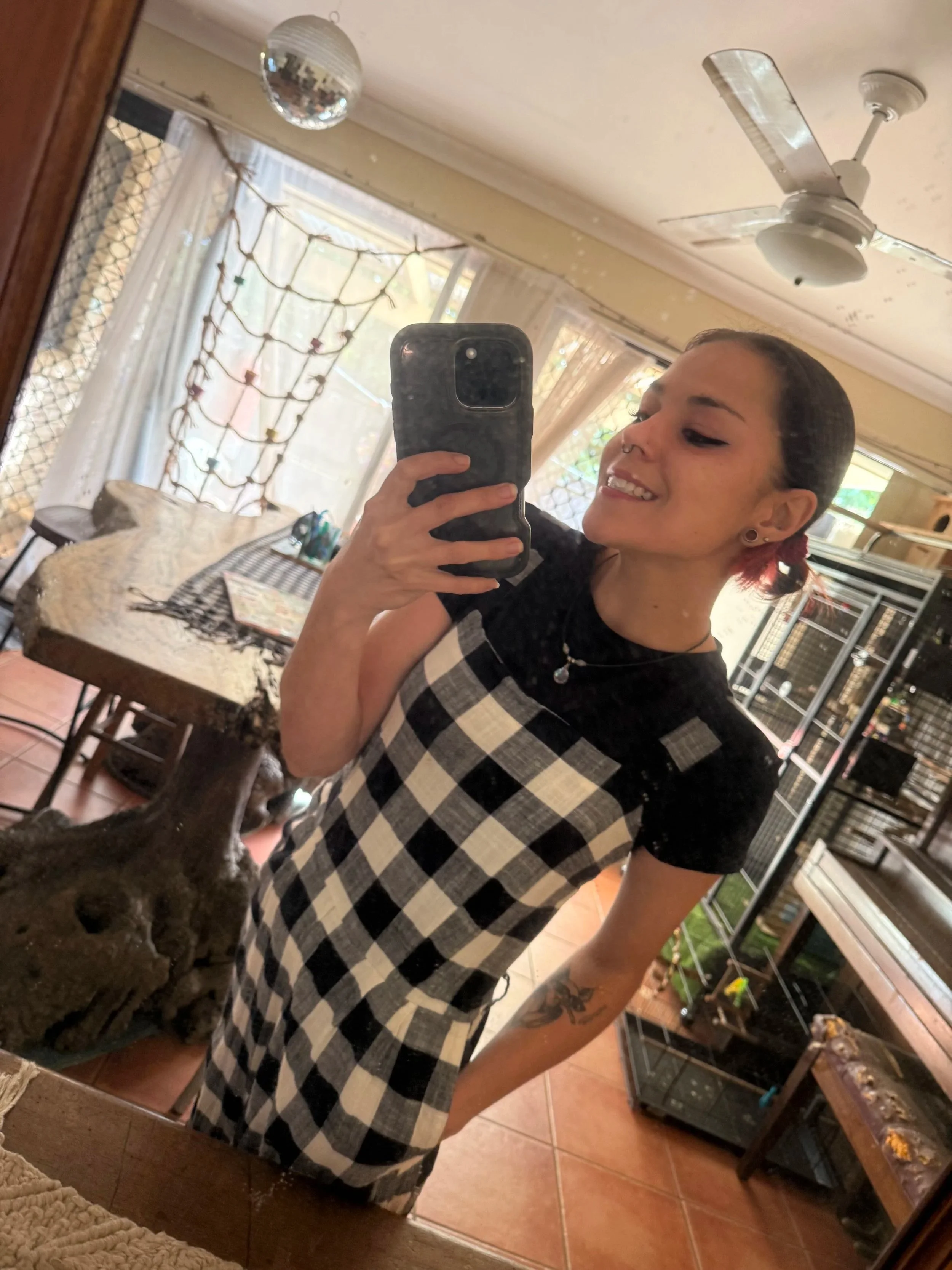 A young woman with dark hair and tattoos on her left arm, wearing a black and white checkered apron, taking a selfie in a mirror in a room with a table, a chandelier, a ceiling fan, and a window with curtains, with a dog visible at her side.