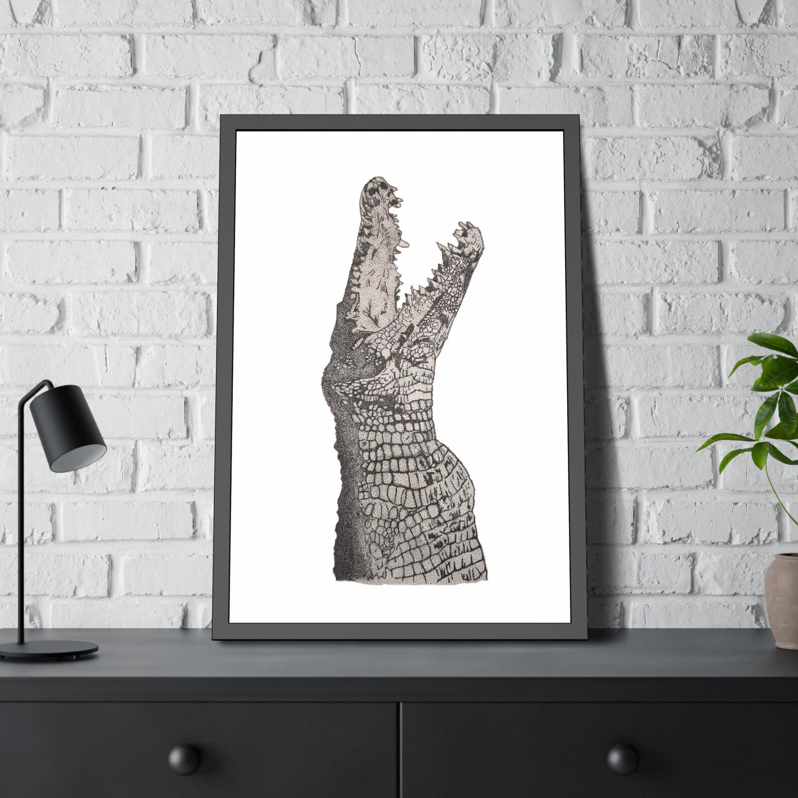 Framed hand drawn stippling illustration of a saltwater crocodile, displayed on a black console table against a white brick wall.