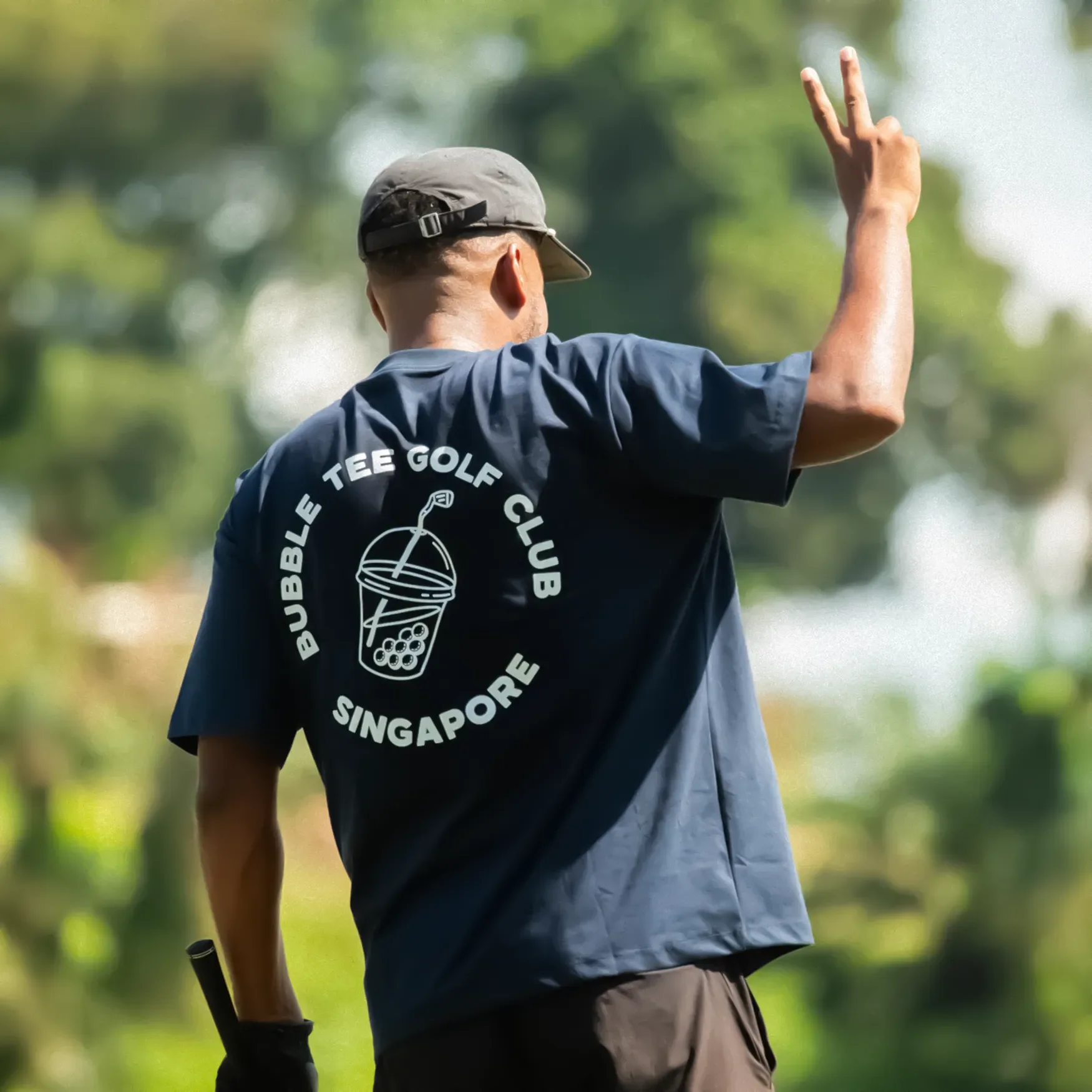 Golfer wearing dark blue Bubble Tee Golf Club t-shirt with large back logo prints, celebrating a shot on a lush Singapore golf course in relaxed premium golf apparel.