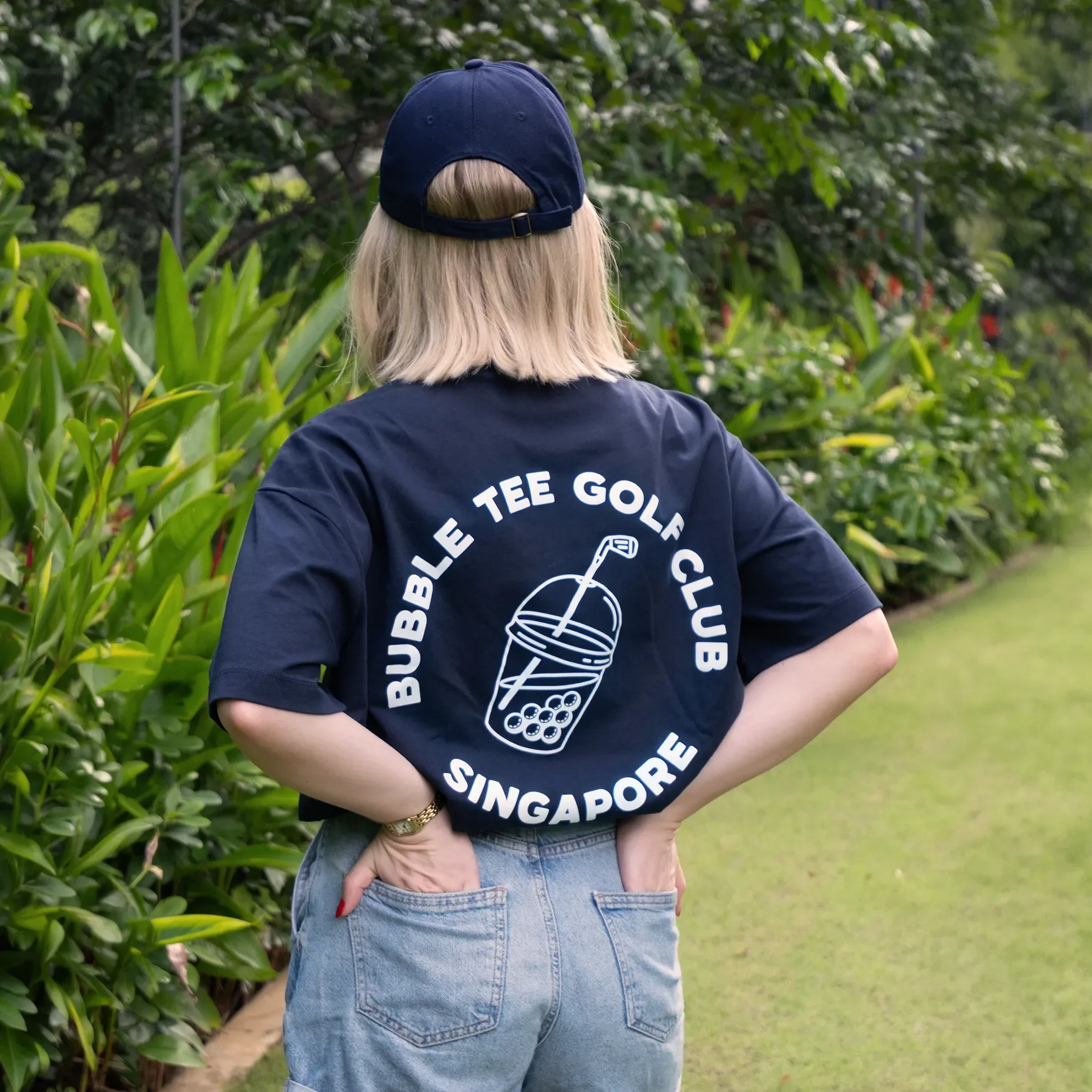 Female golfer wearing navy blue Bubble Tee Golf Club t-shirt with large back logo print, standing on a Singapore golf club fairway in relaxed premium golf apparel.