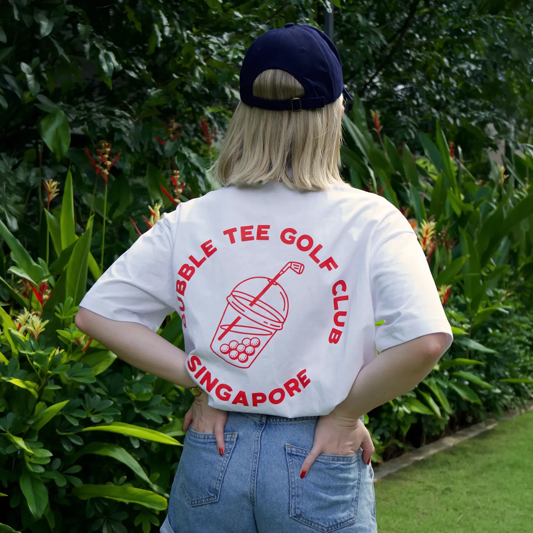 Female golfer wearing white Bubble Tee Golf Club t-shirt with small front logo and large back logo print, standing on Singapore golf course, relaxed premium golf apparel.