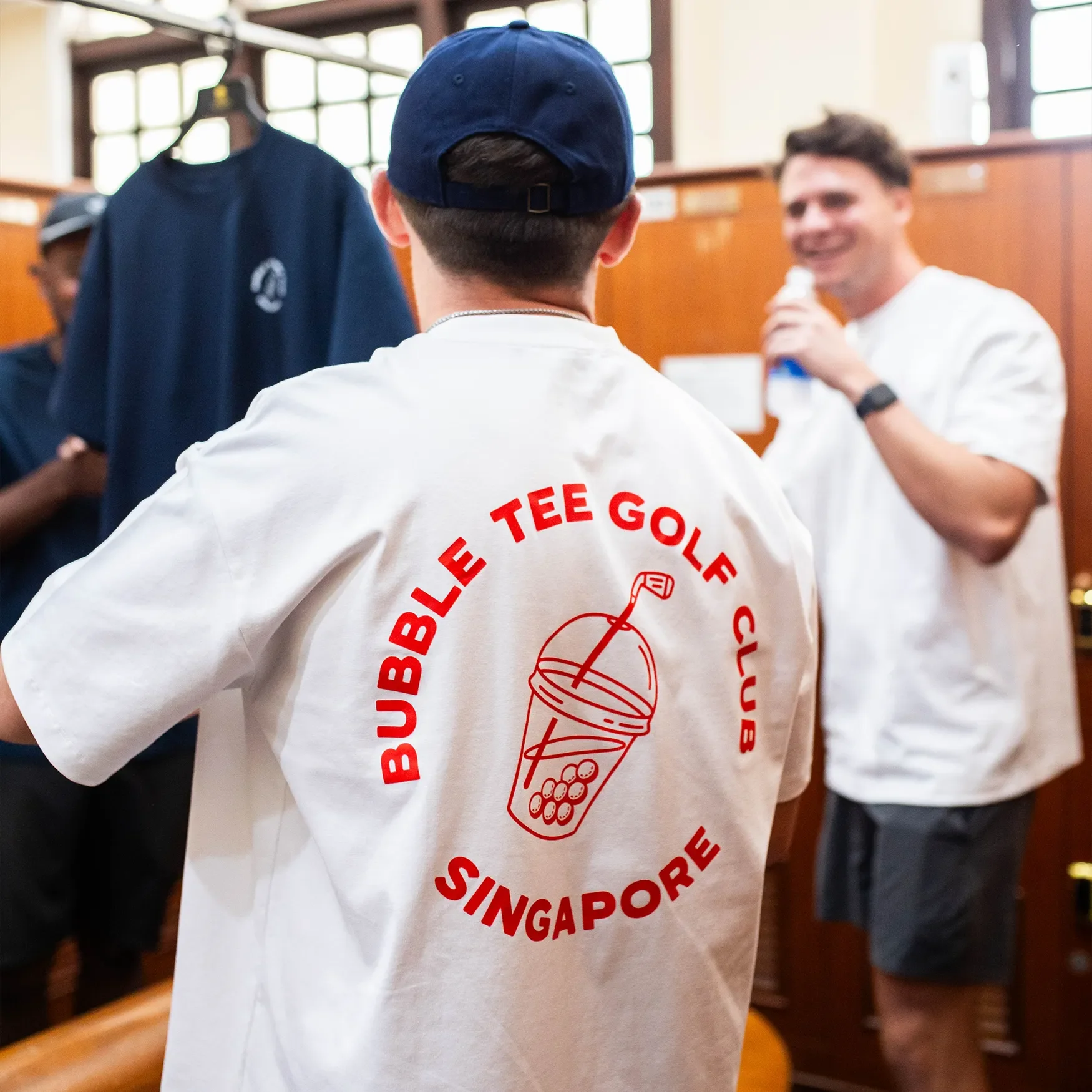 Male golfers wearing white Bubble Tee Golf Club t-shirt with large back logo prints, standing in Singapore golf club changing room, relaxed premium golf apparel.