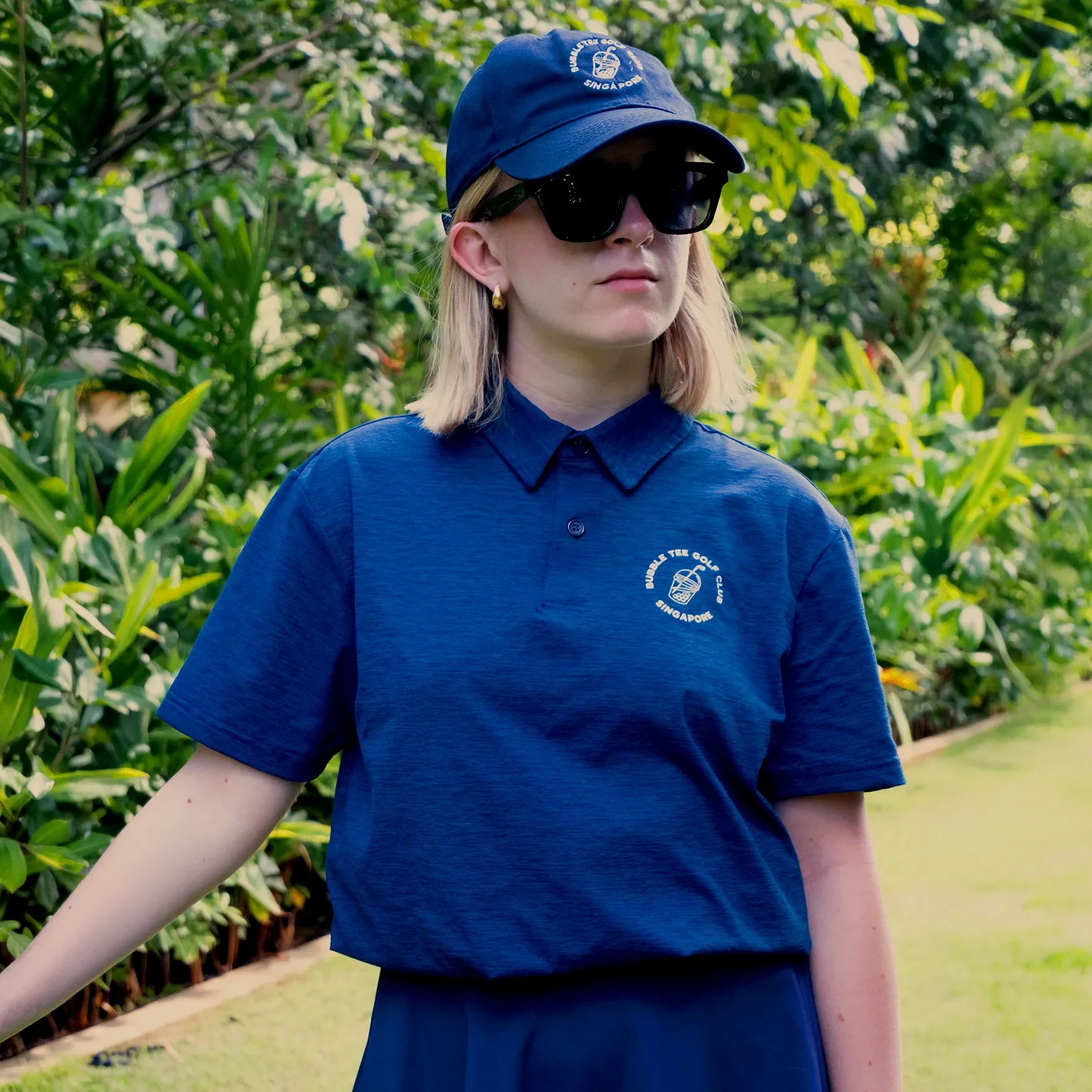 Female golfer waiting to play on a golf course wearing a navy Bubble Tee Golf Club polo shirt with embroidered Singapore logo.