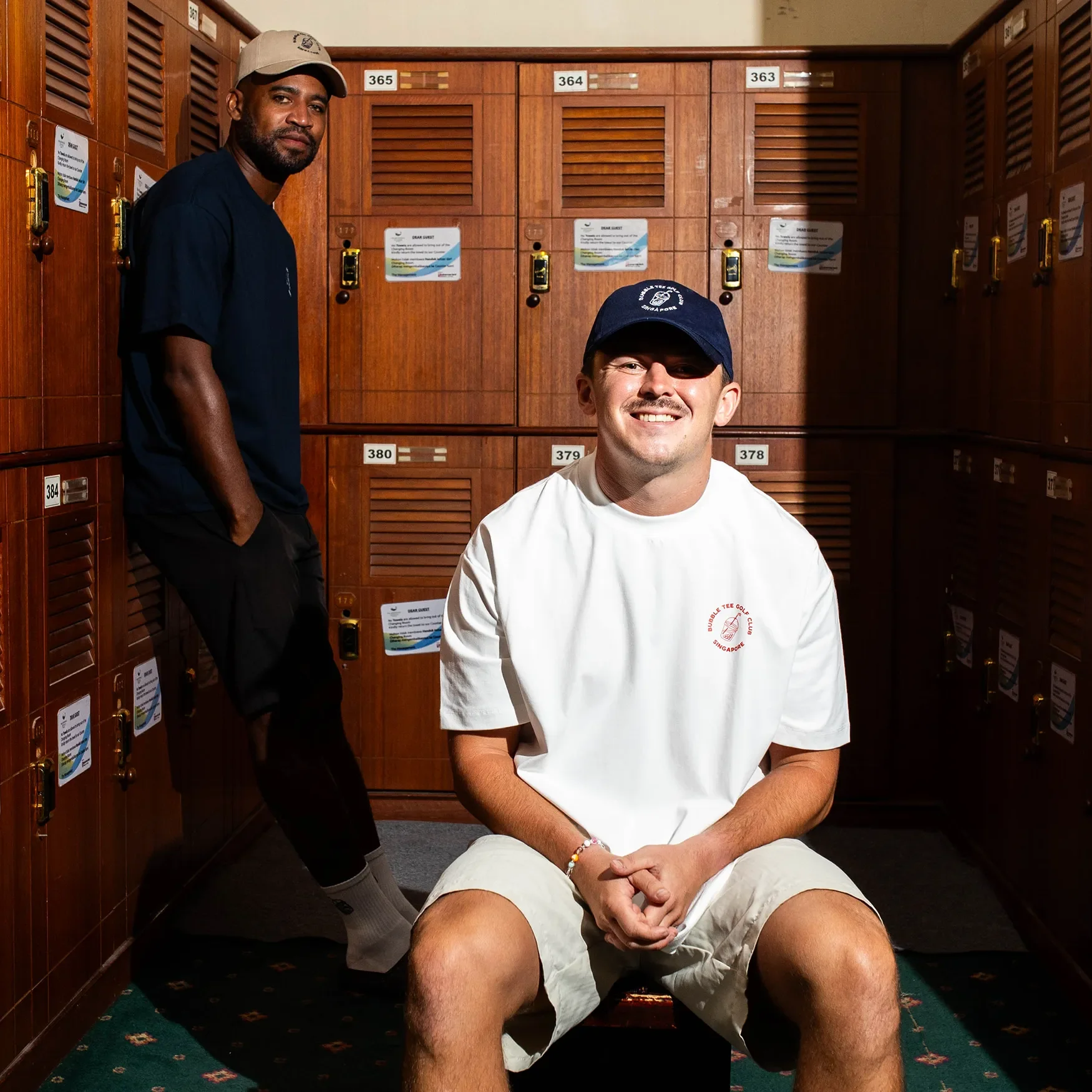 Male golfer wearing white Bubble Tee Golf Club t-shirt with small front logo and large back logo print, sitting in Singapore golf club changing room in relaxed premium golf apparel.