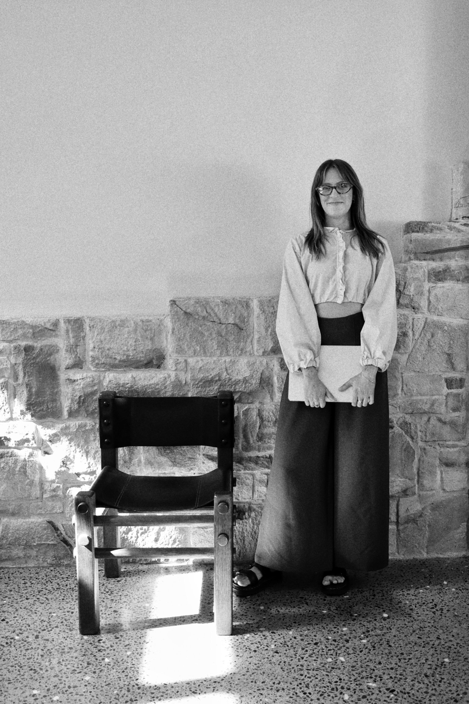 A woman with glasses, wearing a long skirt and sandals, standing indoors against a stone wall background, holding a piece of paper. An empty wooden chair is in the foreground.