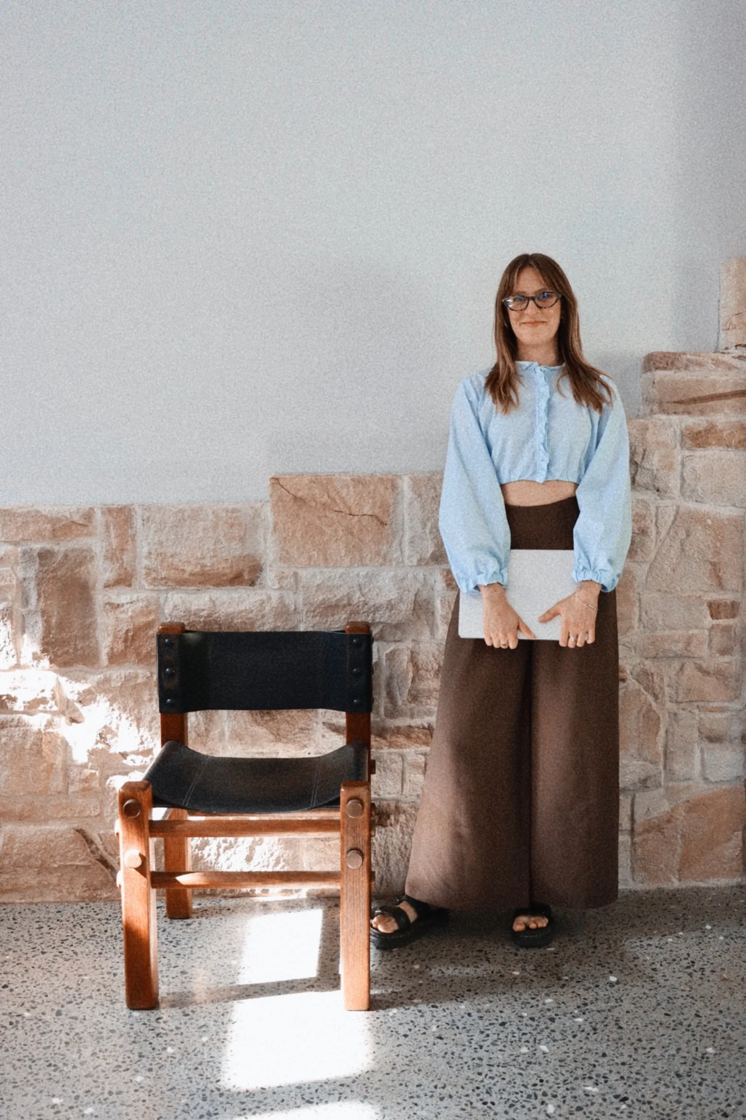 A woman with brown hair, glasses, wearing a light blue blouse and wide-legged pants, standing next to a wooden chair with a black seat, against a brick wall with a white upper section, and holding a white object.