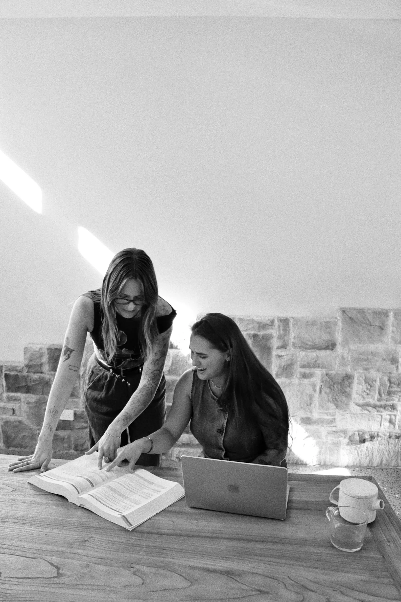 Two women are sitting at a table with open books, a laptop, a mug, and a glass of water. One woman is standing, pointing at the book, while the other is sitting and smiling. They are in a room with a brick wall and a sloped ceiling.