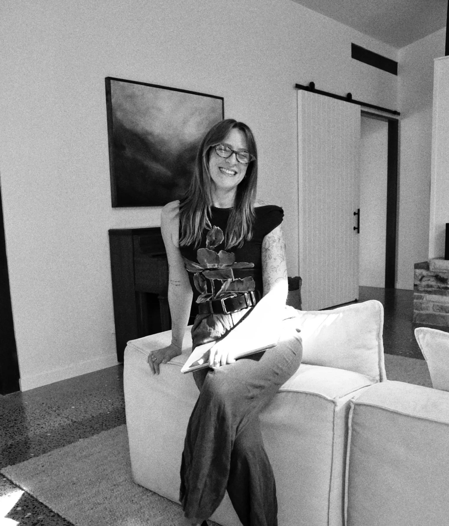 A woman with glasses smiling, sitting on a white armchair with one arm resting on the back and holding a notebook with her other hand, in a living room with a painting behind her and a sliding barn door in the background.