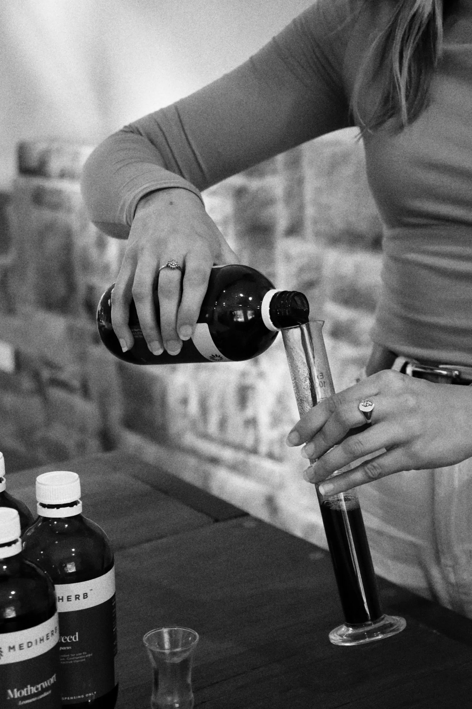 A woman is pouring liquid from a bottle labeled herbal medicine into a test tube held in her other hand. Several bottles and a small glass container are on the table in front of her.