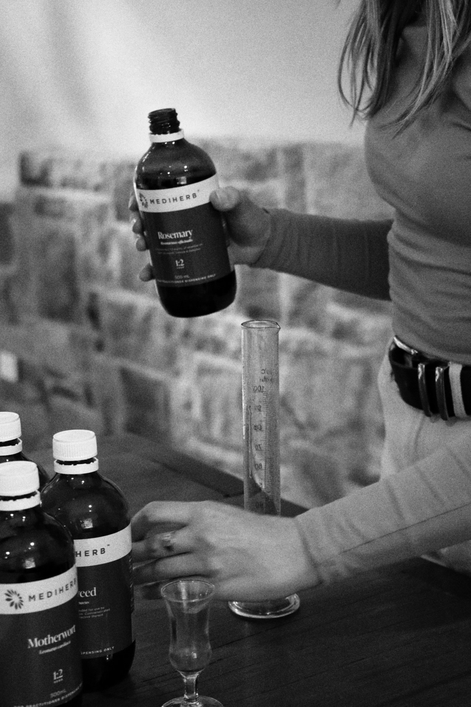 A person holding a bottle ofmediherb herbal supplement while filling a test tube with a liquid. Multiple similar bottles are on the table next to a glass with a small amount of liquid.