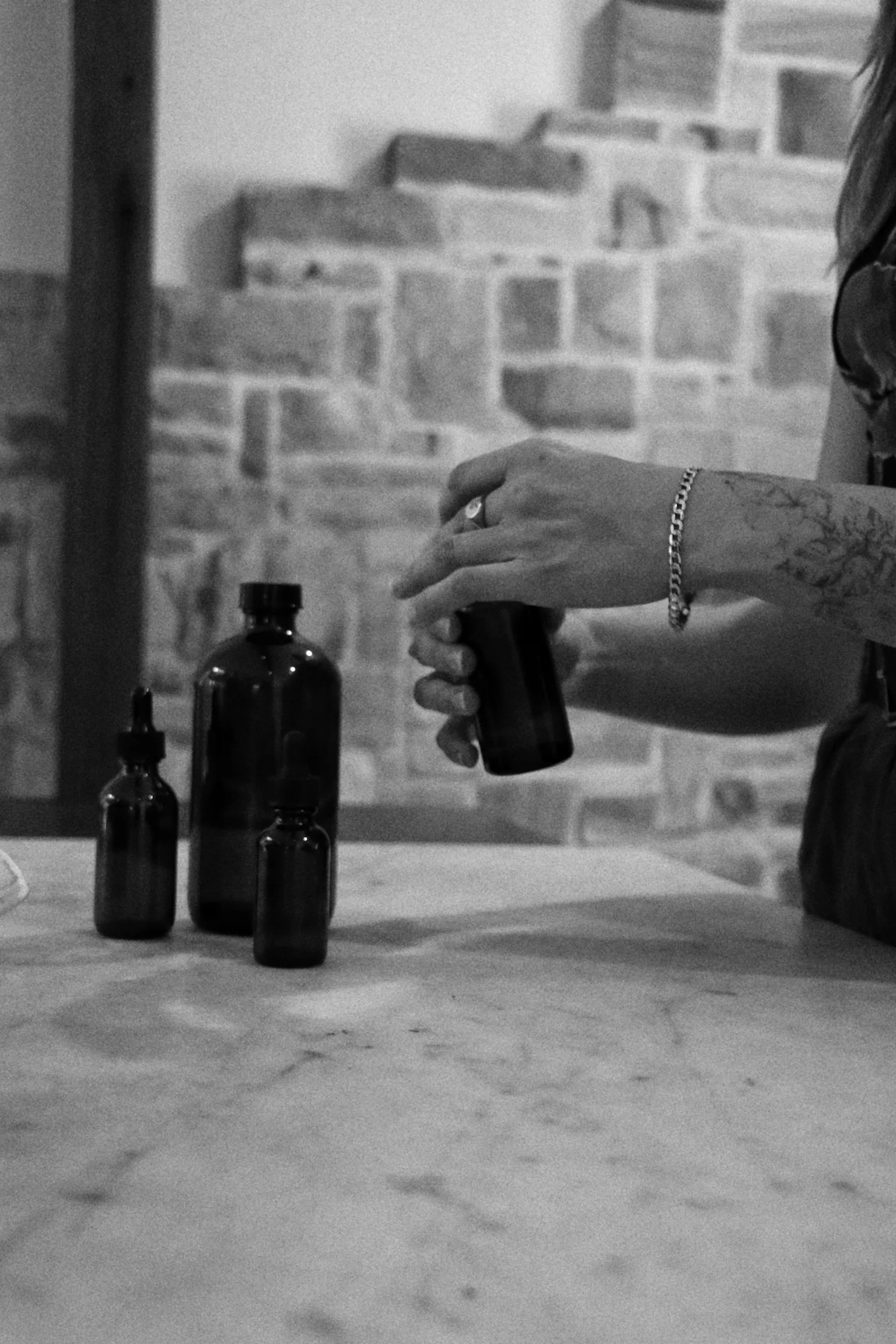 Black and white photo of a person with tattoos on their hand and wrist, wearing a bracelet and a ring, holding a small bottle. Three similar bottles are on a wooden surface in front of them. The background features a brick wall and a wooden beam.