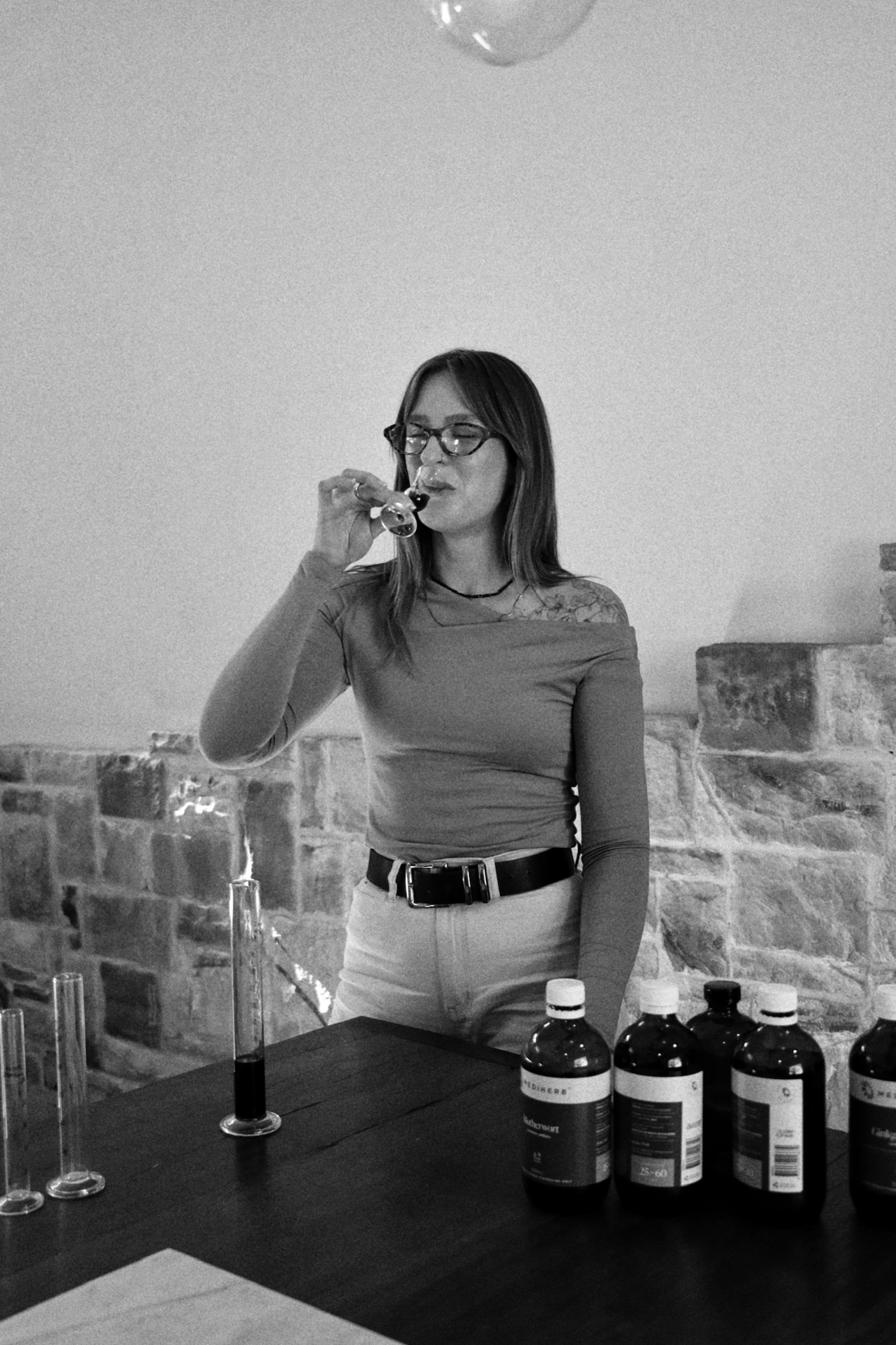 A woman with glasses and long hair is standing at a table with laboratory bottles and test tubes, sparking a glass with a flame, in front of a brick wall.