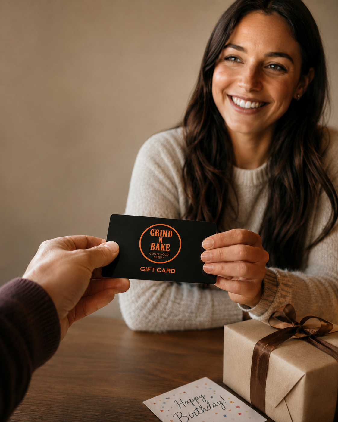 A woman receiving a gift card from a person in a brown sweater, with a wrapped gift and a birthday card on a wooden table.