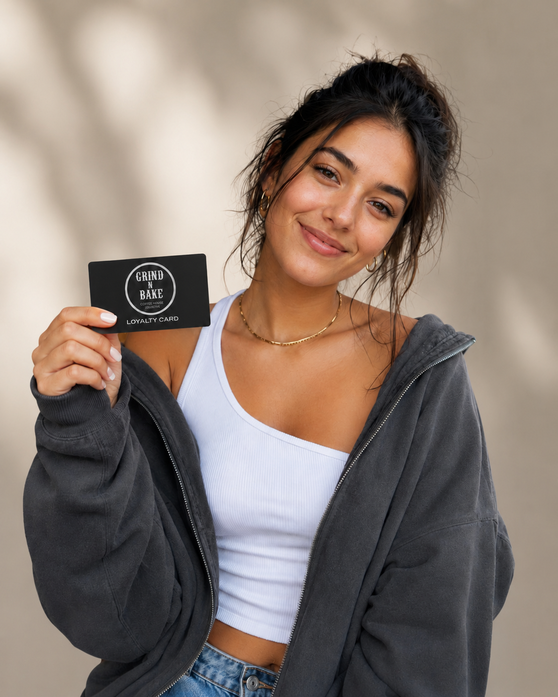 Young woman smiling and holding a black loyalty card that reads 'Grind N Bake Coffee Roasters Collective' against a neutral background. She wears a white tank top, grey hoodie, gold jewelry, and has her hair styled in a messy bun.