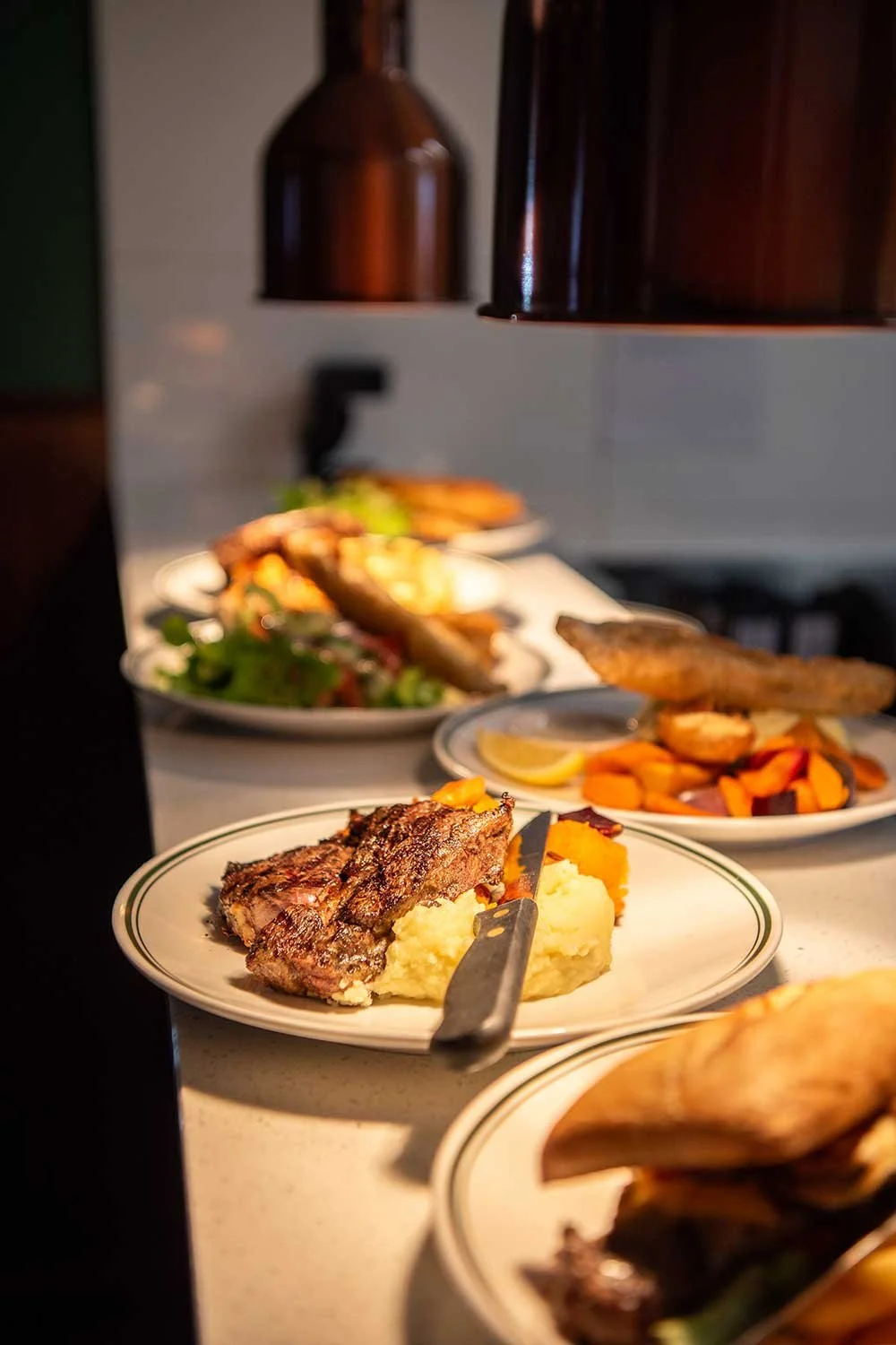 Close-up of a plate of grilled steak with mashed potatoes and roasted vegetables on The Dederang Hotel's kitchen counter, with other plates of food in the background.