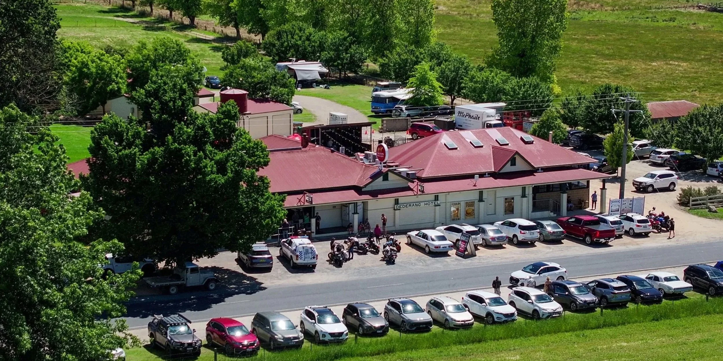 Aerial view of The Dederang Hotel with cars parked in front, surrounded by green trees and grassy areas, and other vehicles and buildings in the background.