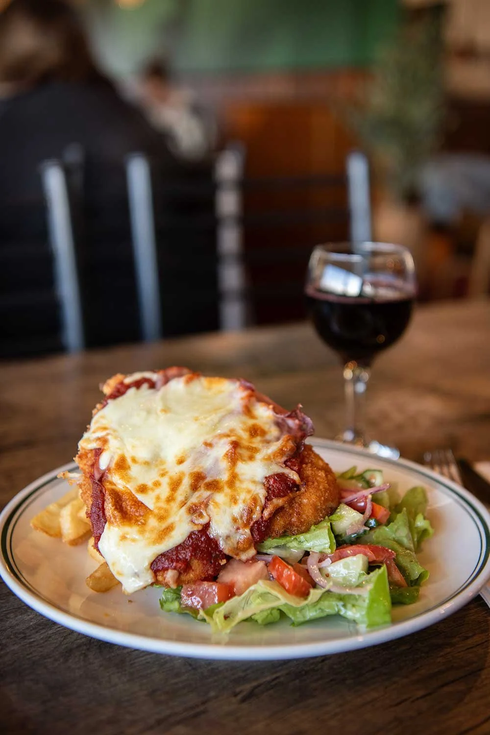 A chicken parmi topped with melted cheese, with a side salad and fries, and a glass of red wine in the background.