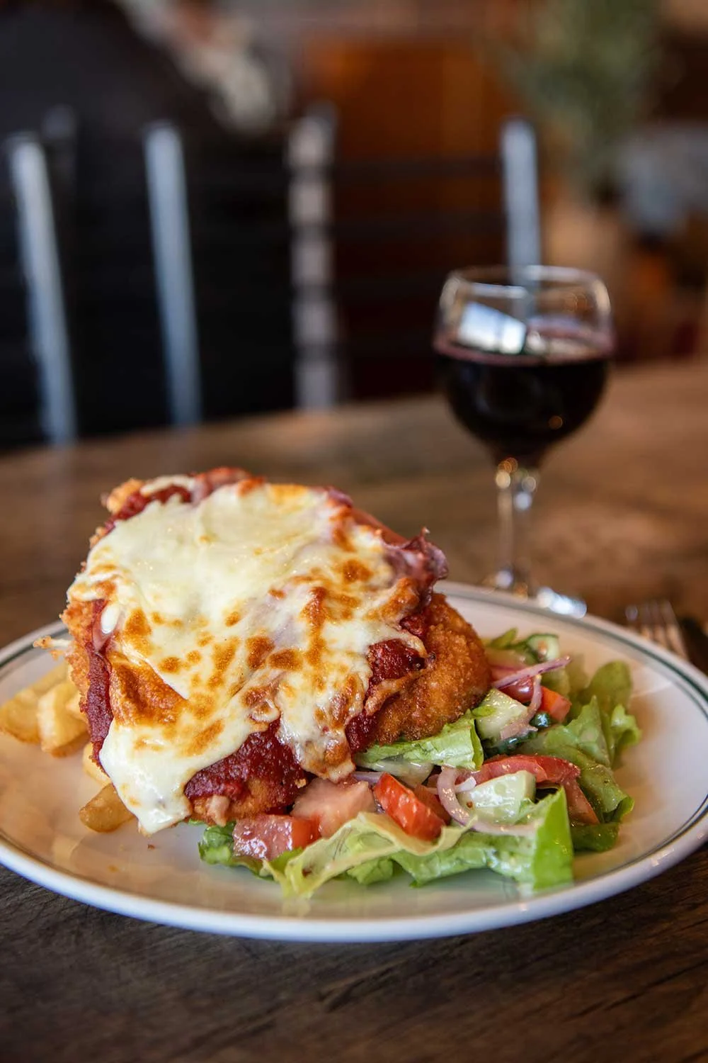 A chicken parmi topped with melted cheese, served with a side salad of lettuce, tomatoes and onions, with a glass of red wine in the background.