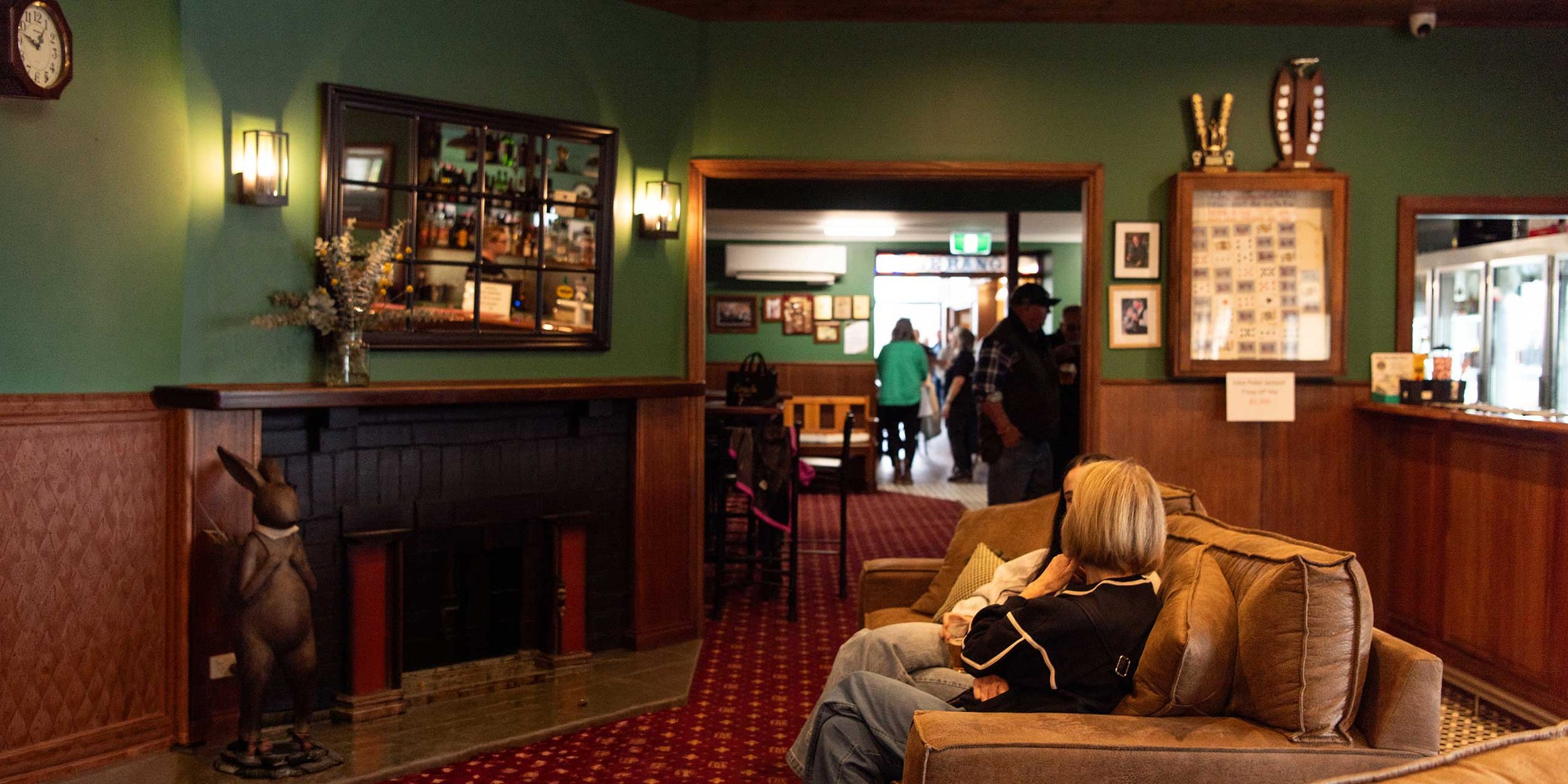 Inside the main entrance of The Dederang Hotel, featuring green walls, wooden accents, framed pictures and a fireplace. People are sitting on a couch and standing, with a view into the pavilion area where more customers are gathered.